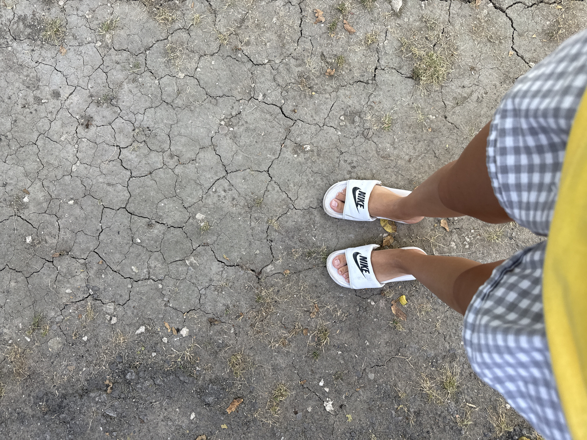 The image shows a pair of feet wearing white Nike slides, standing on cracked, dry earth with sparse dry grass and scattered leaves. The person is wearing a plaid skirt and a yellow top. The scene is outdoors, with the ground suggesting a natural or unpaved area. The location is identified as Basarabeasca, Moldova.