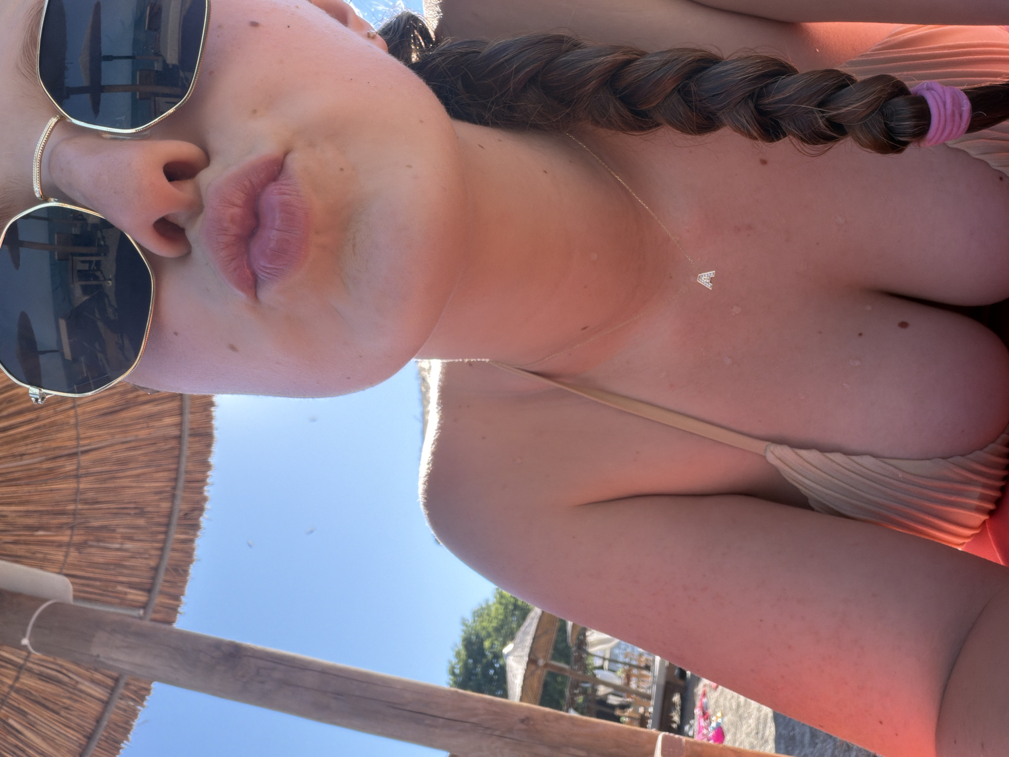 The image is a close-up, low-angle shot of a young woman on a beach. The sun is bright, indicating a clear day, likely midday or afternoon. The woman is wearing sunglasses and a beige bikini. Her hair is styled in a braid and falls over her shoulder, with a pink elastic band securing the end. A delicate, diamond-encrusted letter 'A' necklace is visible on her chest. The background shows a thatched beach umbrella, wooden beams, and a glimpse of trees and buildings, suggesting a resort or beachside location in Burgas, Bulgaria. The overall mood appears relaxed and summery, with the woman perhaps posing for a selfie or enjoying a day by the water.