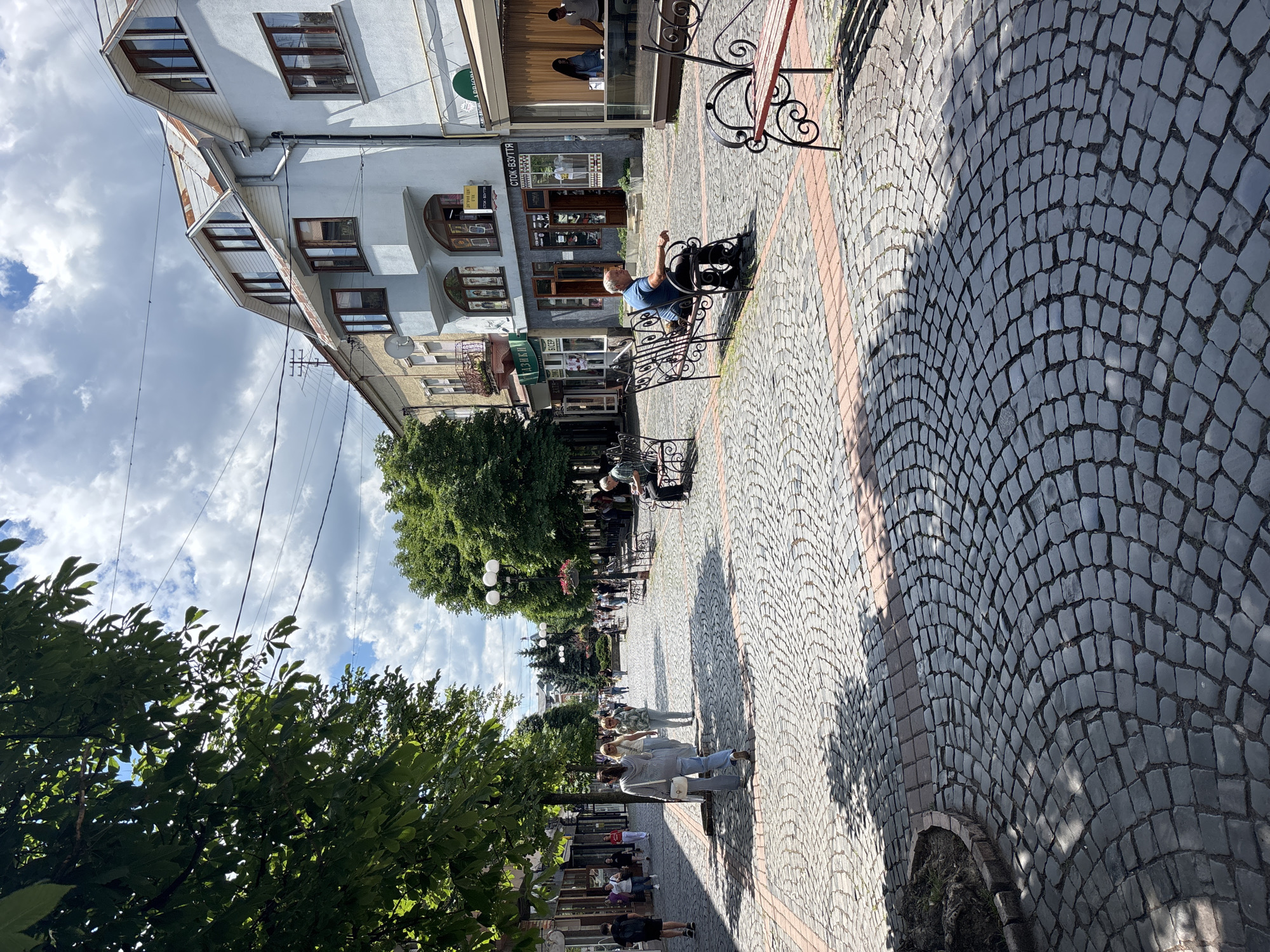 A sunny day in an urban pedestrian zone paved with cobblestones. Lush green trees line both sides of the walkway, casting dappled shadows on the ground. Several people are strolling along the path, while others are seated on wrought-iron benches, enjoying the weather. Buildings with traditional architecture flank the street, some featuring shops with open doorways and windows. The sky above is a mix of blue and white clouds.