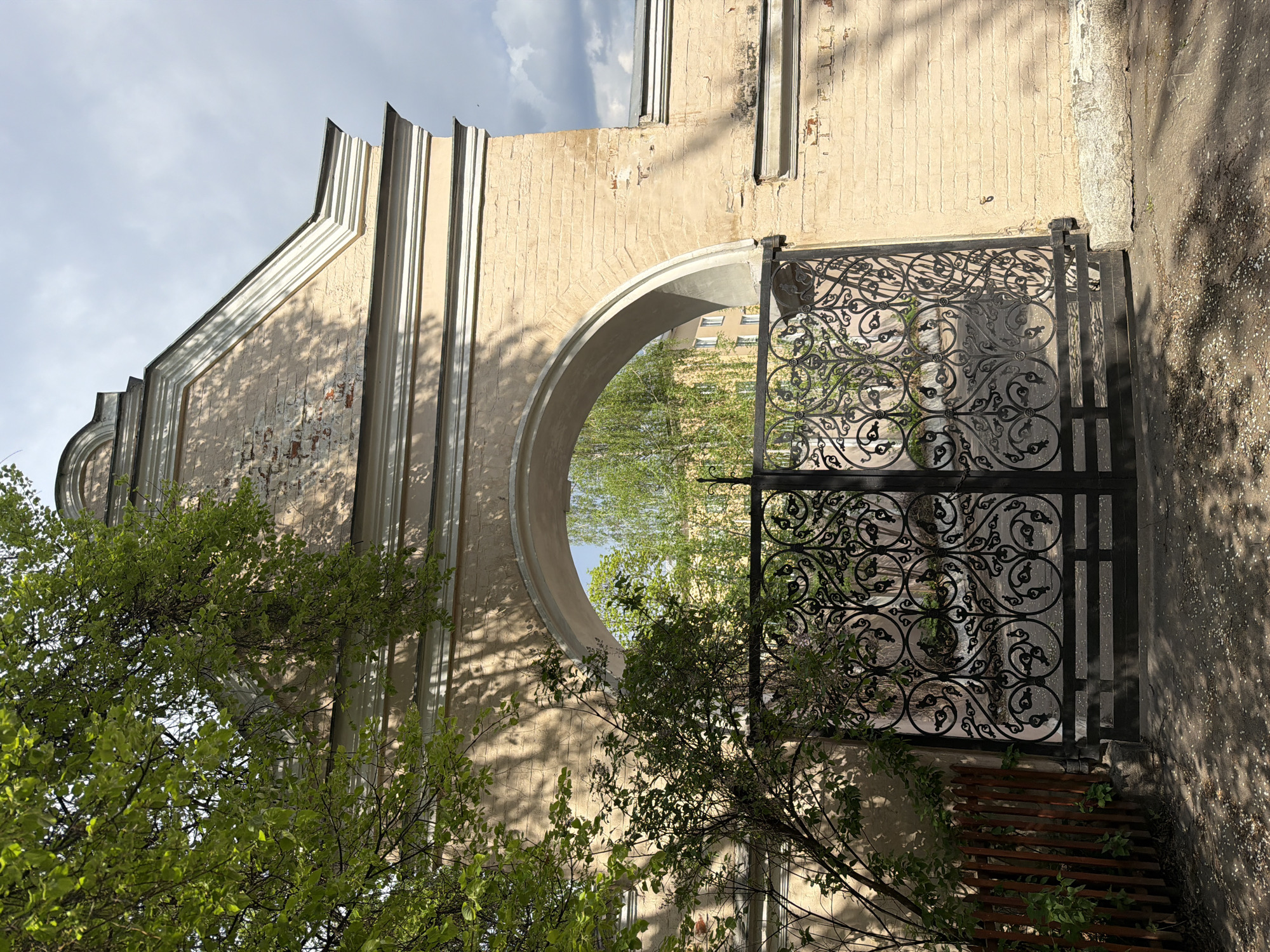 The image shows a stone archway with an ornate black iron gate in Moscow, Russia. The archway is part of an old building with a beige facade and neoclassical architectural details. The paint is peeling and chipped, revealing the brick underneath. The gate is intricately designed with circular and floral patterns. Lush green trees obscure the view behind the gate. The scene is bathed in sunlight, creating shadows on the archway and ground. A section of a wooden bench is visible on the lower right. The sky is partially visible through the arch, with some clouds.