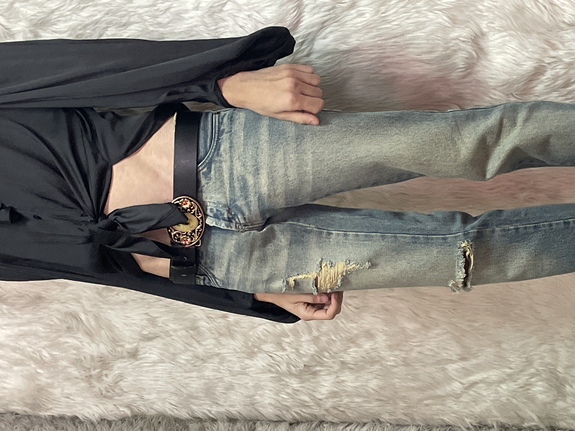 The image shows a person lying on a light-colored, textured surface, possibly a fluffy blanket or rug. The person is wearing light blue, distressed jeans with rips at the knees and a black leather belt with a decorative buckle. They are also wearing a black, long-sleeved top that is tied at the front, exposing a portion of their torso. The hands are resting on the jeans, one near the waist and the other near the knee. The background is indistinct but appears to be the same textured surface. The scene appears to be indoors, possibly in a residential setting. There is no evidence in the image to determine the location in Buenos Aires, Argentina or any other specific geographic location.
