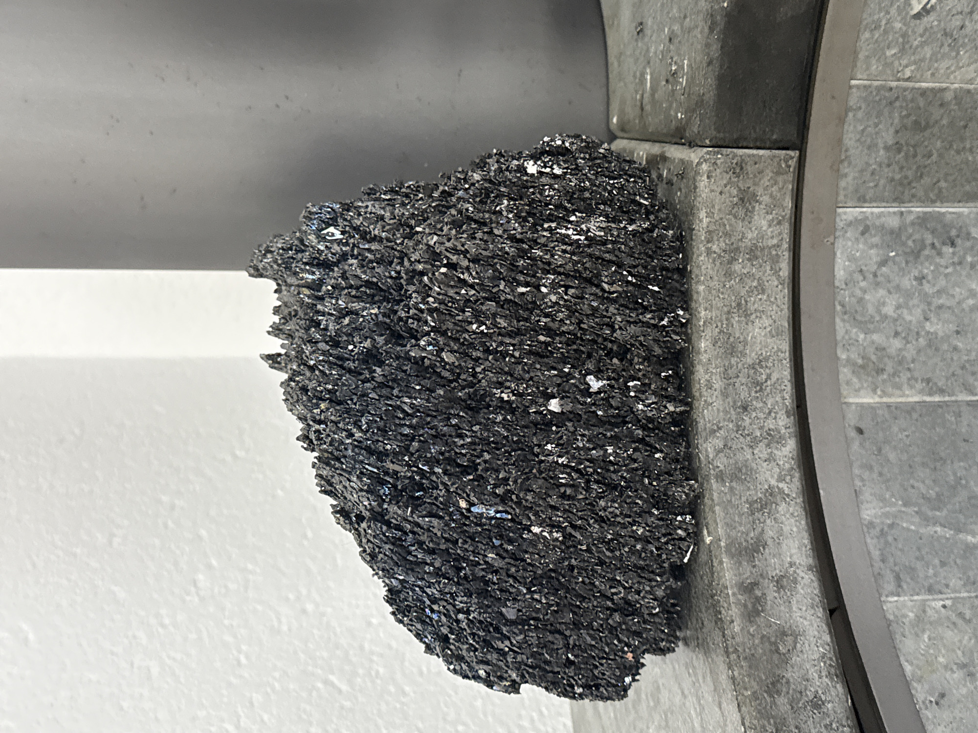 The image shows a dark, crystalline mineral sample resting on a gray, textured surface. The mineral has a rough, multifaceted texture and appears to be composed of many small, sharp crystals. Some of the crystals exhibit a subtle iridescent sheen, reflecting hints of blue and white. The background consists of a plain white wall on the left and a large, cylindrical gray object on the right. The mineral is positioned on a ledge or surface made of segmented gray tiles. Below this ledge, a curved metallic trim separates it from a tiled floor with a pattern of light gray squares. There are no people or any signs of activity depicted in the image. The scene appears to be indoors, possibly in a display or laboratory setting. The image does not provide any information to place the scene in Ulmen, Germany.