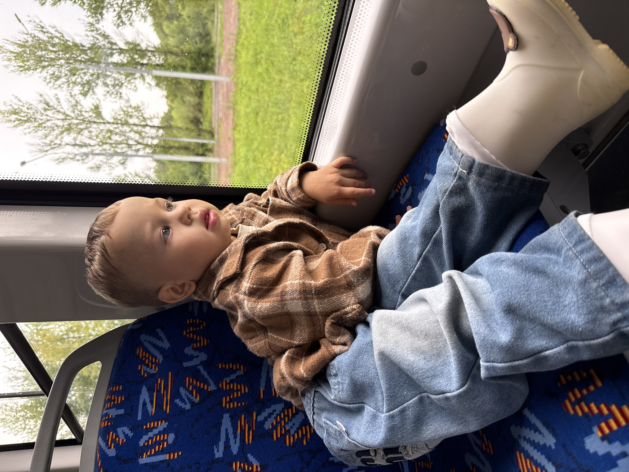 A young Caucasian child, appearing to be around two years old, sits on a blue patterned seat in what appears to be a train or bus. The child is wearing a brown plaid shirt, light blue denim jeans, and white boots. The child's hair is light brown and appears slightly damp. They are looking out a window to the right of the frame, with their left arm resting on the windowsill. Outside the window, green grass and trees are visible under an overcast sky. The interior of the vehicle shows a grey handrail above the seat and the patterned upholstery of other seats. The image was taken in Rusanovo, Russia.