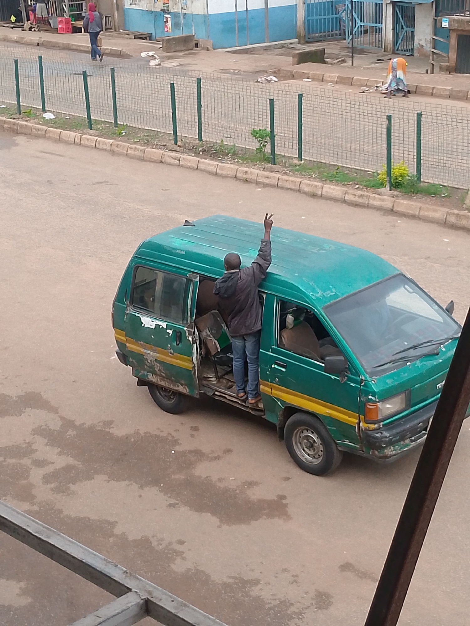 An overhead medium shot captures a vibrant green and yellow public transport van on a paved street in Bukuru, Nigeria. The van, which shows signs of wear and tear with chipped paint, has its passenger door open, revealing a man standing in the doorway, his arm raised with two fingers in a peace sign. He wears a dark jacket and blue jeans. The street appears to be wet in patches, suggesting recent rain. In the background, a metal fence separates the street from a sidewalk lined with businesses. A few pedestrians are visible on the sidewalk, one wearing a red headscarf and blue jeans, and another in a colorful dress and orange headwrap. The overall scene depicts a busy urban environment.