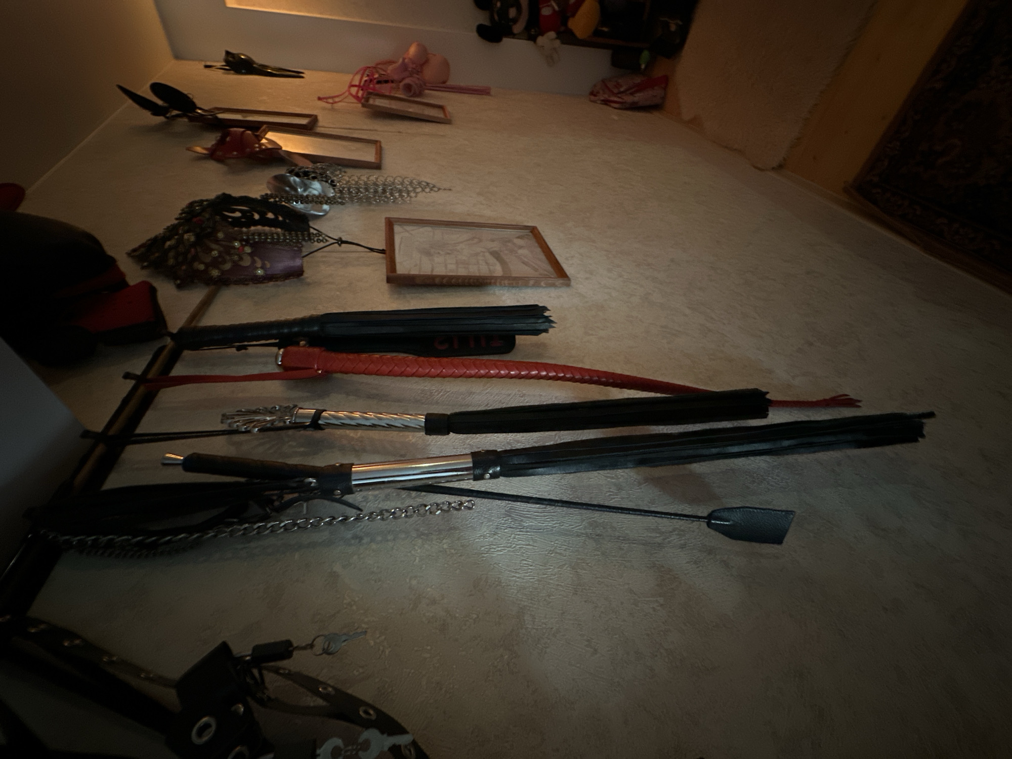 The image depicts an assortment of items scattered on a surface, possibly a floor or table, with the perspective angled upward. The objects include various types of whips, a mask, and what appear to be framed pictures. There are also some restraints. The presence of these items suggests a possible scene related to BDSM. The location cannot be determined from the image.