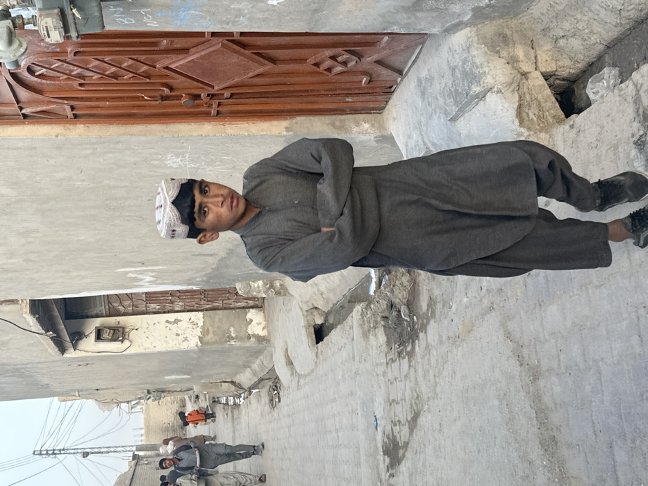 The image depicts a narrow, paved alleyway in Quetta City Tehsil, Pakistan, under daylight conditions. In the foreground, a male individual, appearing approximately 10-14 years old, stands with his arms crossed over his chest, looking directly towards the camera. He wears a dark grey shalwar kameez and black athletic shoes, along with a white and black patterned cap on his head.
To the subject's right, a plain, light-colored building wall features a prominent reddish-brown metal door with a distinct diamond-paneled design. Above the door, a utility meter or box is affixed to the wall. An open concrete drainage channel, containing dark water and debris, runs along the left side of the paved pathway, adjacent to the subject.
Further down the alley, two other male individuals are visible walking away from the camera. One of these individuals carries a white platter or tray. Beyond them, an adult figure in light-colored clothing and a small child in a bright orange garment are also present. The alley is lined with several modest, light-colored buildings exhibiting varying degrees of wear. Numerous electrical wires are visible strung overhead against a hazy, light-colored sky. The ground surface is composed of interlocking pavers, with scattered dirt and refuse, particularly near the drainage channel.