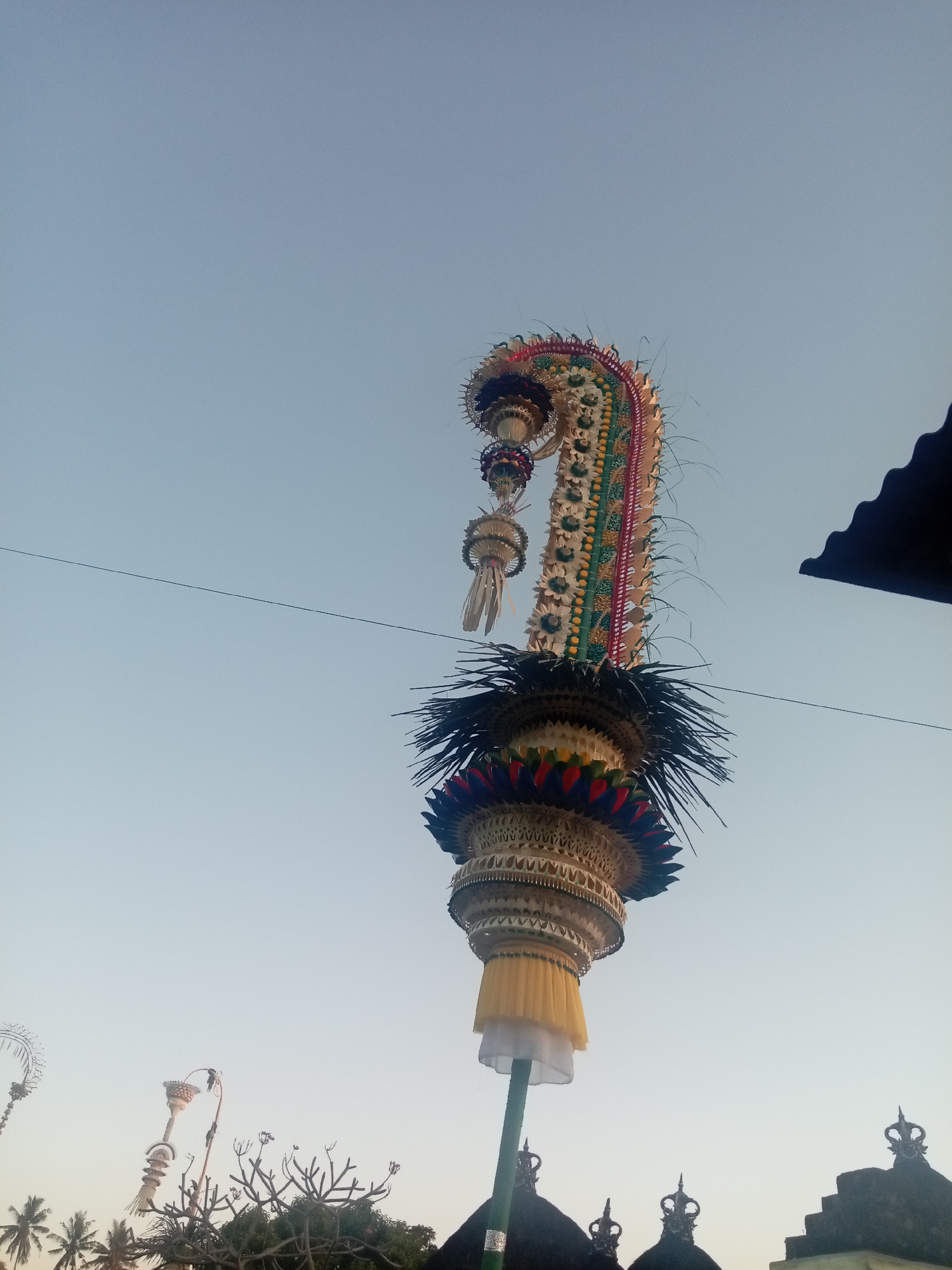 The image shows a tall, ornate religious offering, likely a Penjor, which is a ceremonial bamboo pole, standing against a clear sky. The Penjor is decorated with intricate woven materials, colorful fabrics, and what appear to be natural elements such as leaves and grass. It is topped with a curved, elaborately decorated section that resembles a mythical creature or a symbolic headdress. Below this, layers of woven straw or palm leaves form a tiered structure, culminating in a yellow and white fabric skirt. The Penjor is supported by a green pole. In the background, the dark, sloped roof of a building is visible on the right side. At the bottom left, several other, smaller decorative poles or offerings are partially visible, along with the tops of trees and some architectural elements that appear to be part of a temple or traditional Indonesian structure. The sky is a pale blue, suggesting it is either early morning or late afternoon. The overall scene is typical of ceremonial decorations found in Bali, Indonesia, particularly during religious festivals. Sukawati, Bali is known for its traditional crafts and vibrant religious culture, making this an appropriate setting for such an offering.