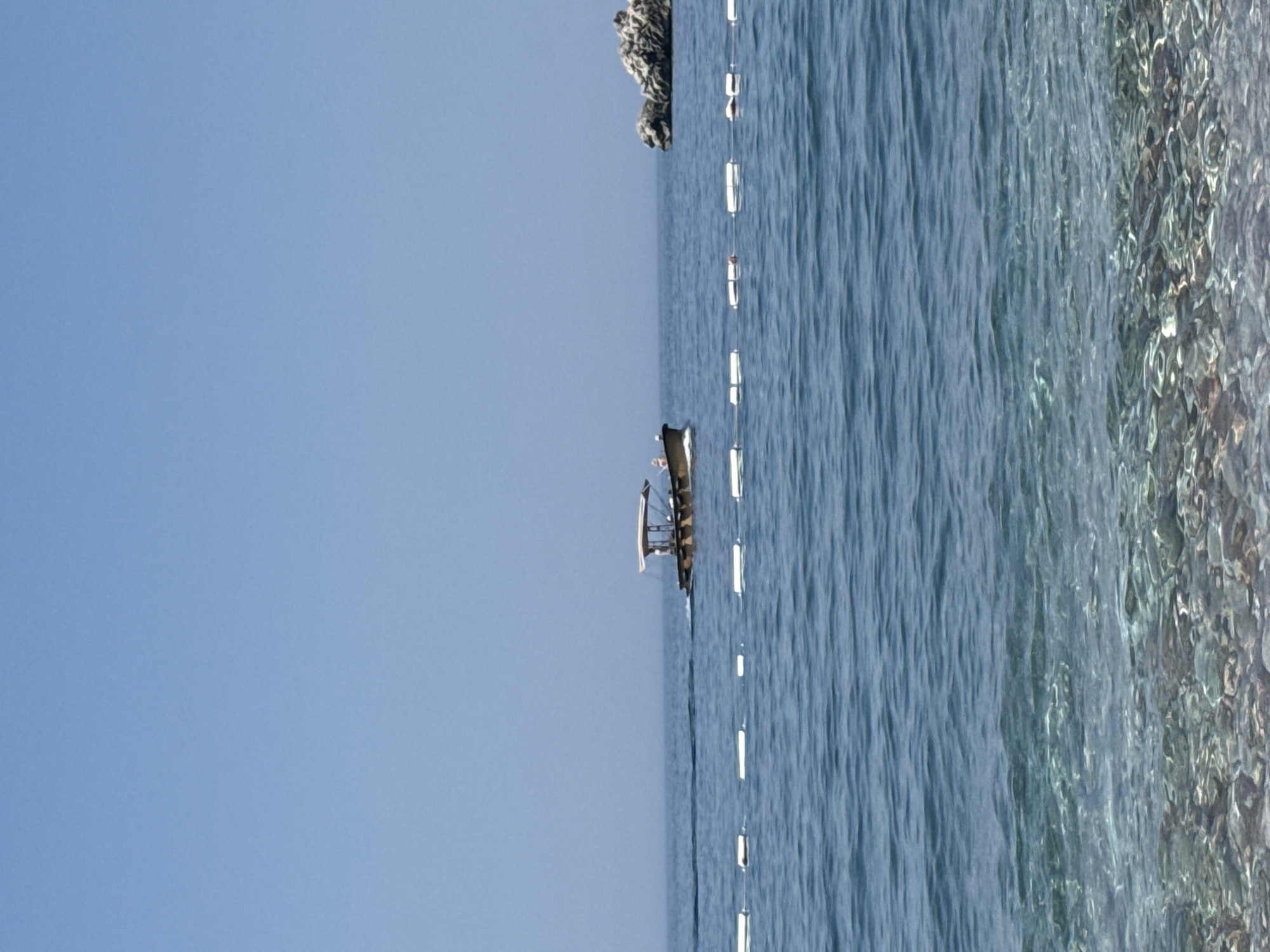 The image depicts a serene seascape with a clear blue sky dominating the left half of the frame. The right side of the image is filled with the calm blue waters of the sea, with ripples indicating a gentle breeze. Along the edge of the water, the seabed is visible through the clear water, showing a textured surface with rocks and pebbles. A string of white buoys stretches across the water, marking a boundary or a swim lane. In the middle ground, a small, dark-colored boat with a canopy is visible, appearing to be stationary. In the upper left background, a rocky outcrop or a small island juts out of the water. The overall impression is one of tranquility and a peaceful day at the beach or sea. The lighting suggests it is daytime, likely late morning or afternoon, with the sun casting a bright, even light. The location appears to be a coastal area, possibly a beach or a harbor, given the presence of buoys and a boat. There is no visible text in the image.