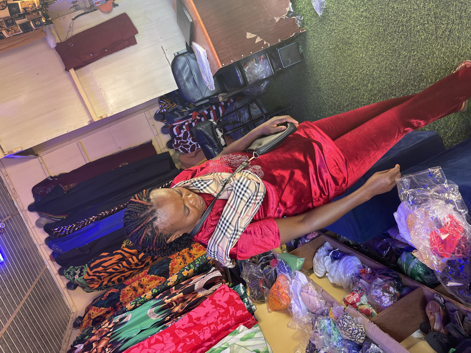 An adult female is depicted standing within a retail clothing establishment in Samaru, Nigeria. She has braided hair with red accents and wears a red velvet short-sleeved tunic with embellished detailing on the chest, along with matching red velvet trousers. A plaid-patterned shoulder bag with a black strap and a small black purse are worn across her body. Her gaze is directed downwards, and her right hand holds a clear plastic bag containing unidentified items.
The environment features a corrugated metal ceiling and light-colored paneled walls. To the left, multi-tiered shelves display numerous items, presumably clothing or textiles, individually packed in clear plastic bags of various colors. Along the back wall, several garments, including dresses and kaftans in diverse patterns (e.g., floral, abstract, animal print) and solid colors (e.g., green, red, orange, blue, black), are hanging on display racks. To the right, a wooden counter is visible, on which rests a black bag and a dark, flat object resembling a laptop or notebook. The floor is covered with green artificial turf. Overhead lighting fixtures are present, with a blue light visible near the ceiling.