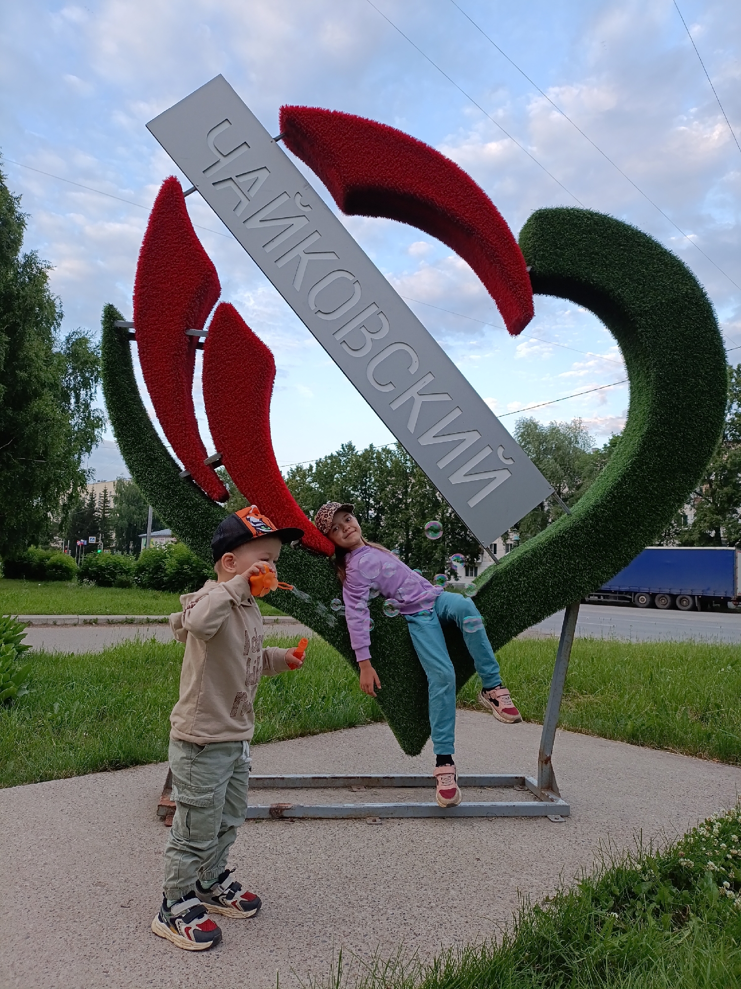 Two children are posed in front of a large heart-shaped sculpture with the Cyrillic inscription "ЧАЙКОВСКИЙ" (Chaikovsky) on a metal plate. The sculpture is made of green artificial grass and red decorative elements. On the left, a young boy wearing a beige hoodie, green cargo pants, a black and orange baseball cap, and colorful sneakers is blowing bubbles. He holds an orange bubble wand. On the right, a girl wearing a light purple hoodie, blue pants, a leopard-print baseball cap, and pink sneakers is leaning back on the sculpture, with her legs bent. Several soap bubbles are floating in the air between the children and the sculpture. The background shows a street with a blue truck, trees, and a cloudy sky. The sculpture is located outdoors, likely in a public park or square in the city of Chaikovsky, Russia, as indicated by the inscription.