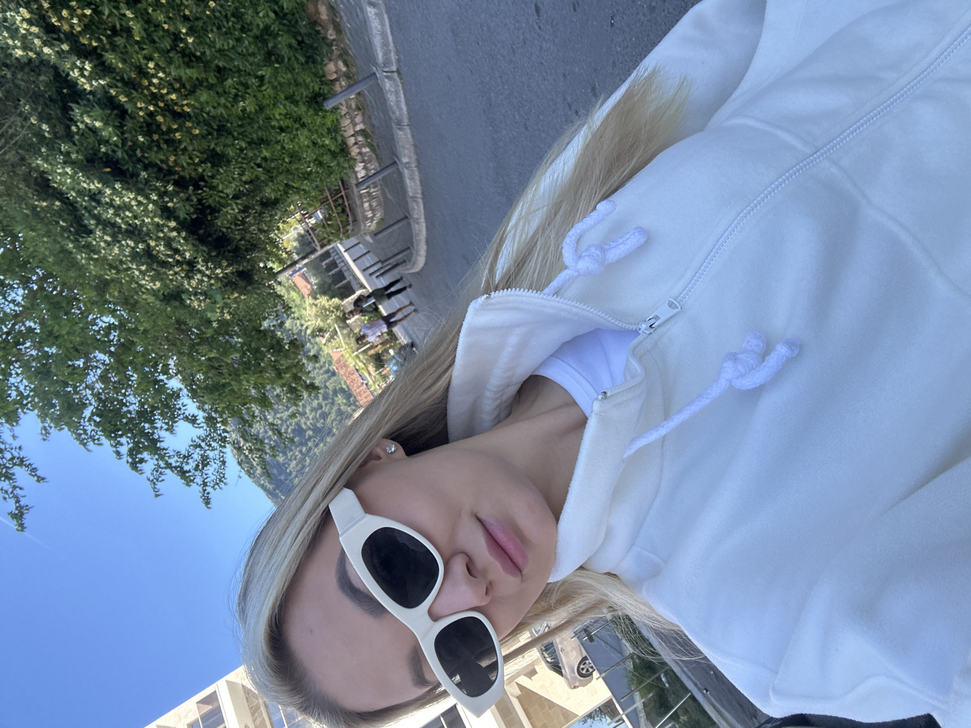 A blonde woman wearing white sunglasses and a white hoodie takes a selfie. She stands in Budva, Montenegro, near a tree-lined street with a metal fence on the left, and a building in the background.