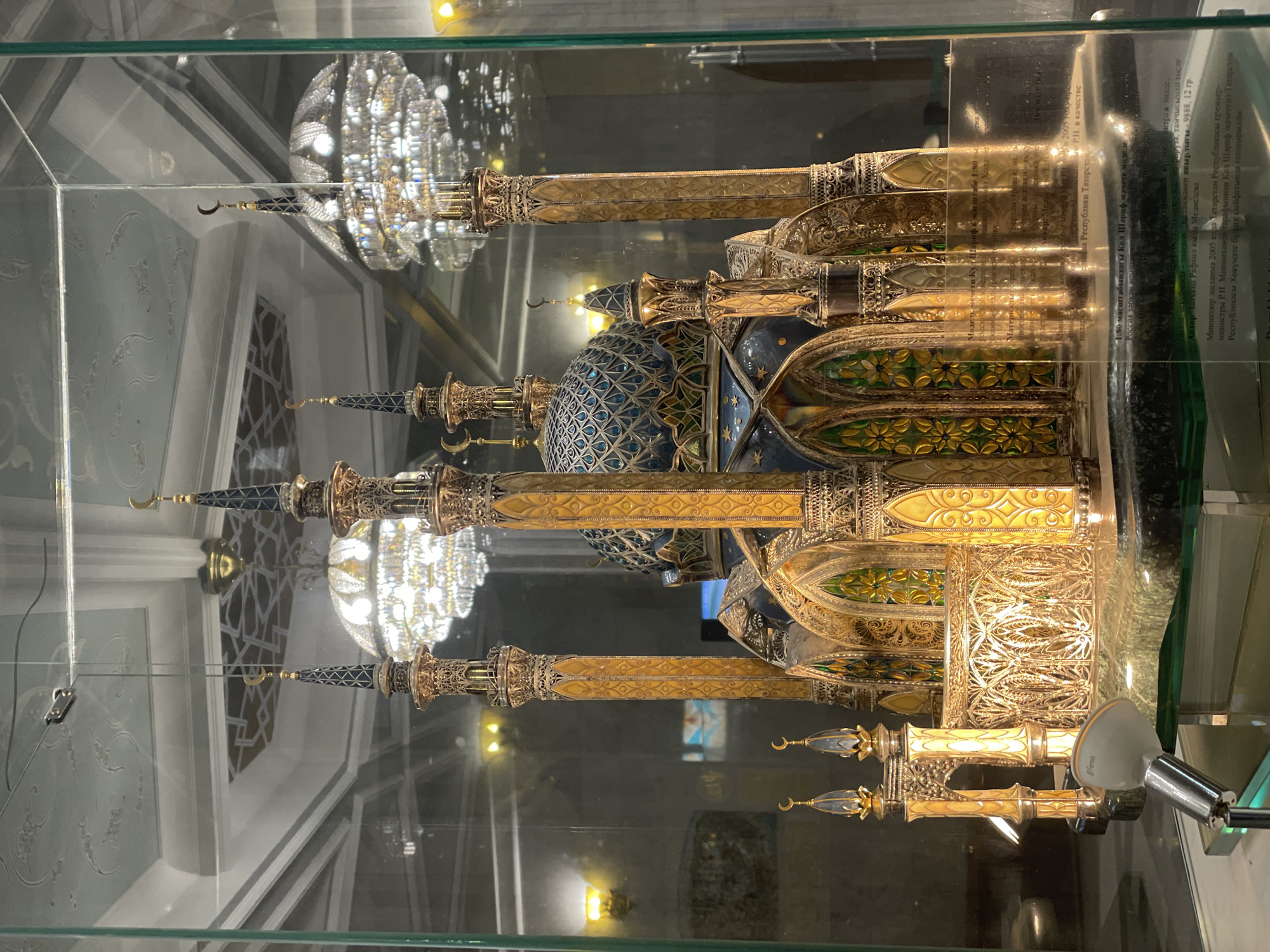 An intricately detailed model of the Kol Sharif Mosque is displayed within a clear glass case. The model, depicted at a 1:80 scale, features multiple minarets topped with crescent moon finials and prominent domes. It is crafted with elaborate gold and silver-colored metalwork, intricate filigree, and vibrant enamel detailing in blue, green, and yellow.
A small "Faron" branded spotlight from the lower right corner of the display case illuminates the miniature. Reflections on the glass reveal parts of the surrounding interior, including ornate chandeliers and decorative wall patterns, suggesting a museum or exhibition setting.
A plaque inside the case provides information in Russian and Tatar. It identifies the model as the Kol Sharif Mosque, weighing 9.888 kg, and crafted with materials like artistic stained glass enamel, turquoise, and serpentine. The author is Tatiana Rafail kyzy Medvedeva. The plaque also notes that the miniature was created in 2005 and presented to the Kol Sharif Mosque by R.N. Minnikhanov, then Prime Minister of the Republic of Tatarstan, as a gift from the Tatarstan Government.