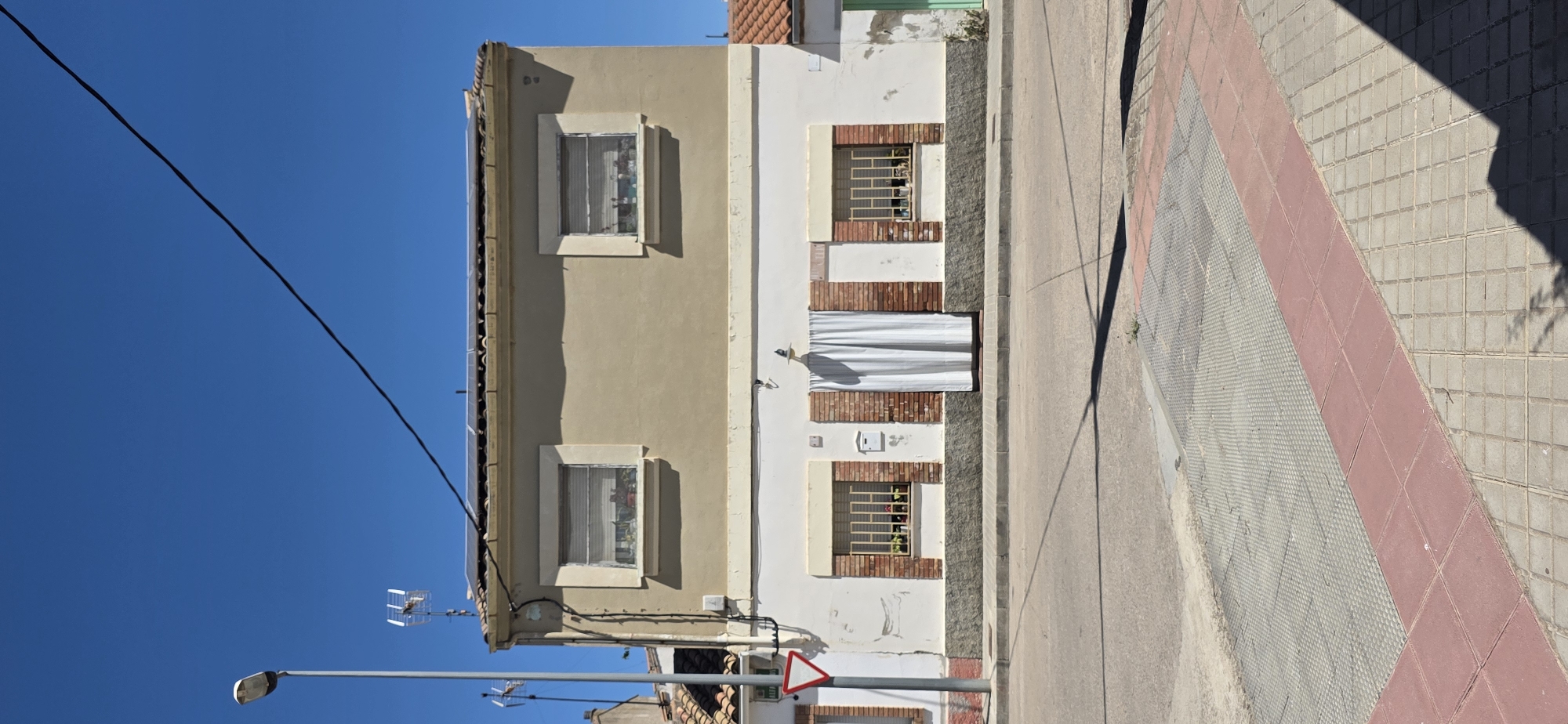 The image depicts a residential building on a street in Huesca, Spain. The building is two stories high with a light brown upper facade and a white lower facade. The upper facade features two evenly spaced windows with horizontal blinds. The lower facade has two windows with barred fronts and plants in the window boxes. A white curtain hangs in the center of the lower facade, covering what appears to be a doorway. The building has a tiled roof and solar panels are visible on top.
In front of the building, there is a paved sidewalk and a street. A traffic sign shaped like a red triangle with a white center is located on a pole to the left of the building. A streetlamp stands to the far left. The sky is clear and blue. The scene appears to be under direct sunlight, casting shadows on the street and sidewalk.