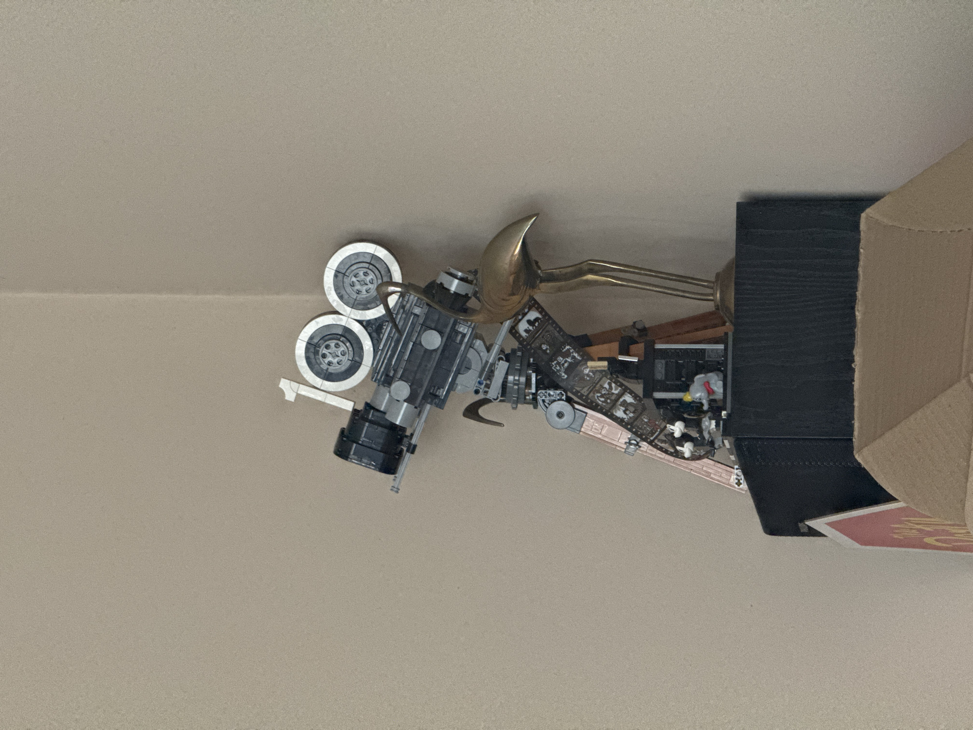 A medium shot of a vintage-style camera rests on a light brown surface. The camera is partially disassembled, exposing various mechanical components and film spools. Next to the camera is a gold-colored horn and a dark wooden box. Adjacent to the box is a partially visible brown cardboard box. The setting appears to be indoors, with a uniform, light-colored background suggesting a tabletop or similar flat surface. No people are visible in the image. The location is not discernible from the visual information provided.