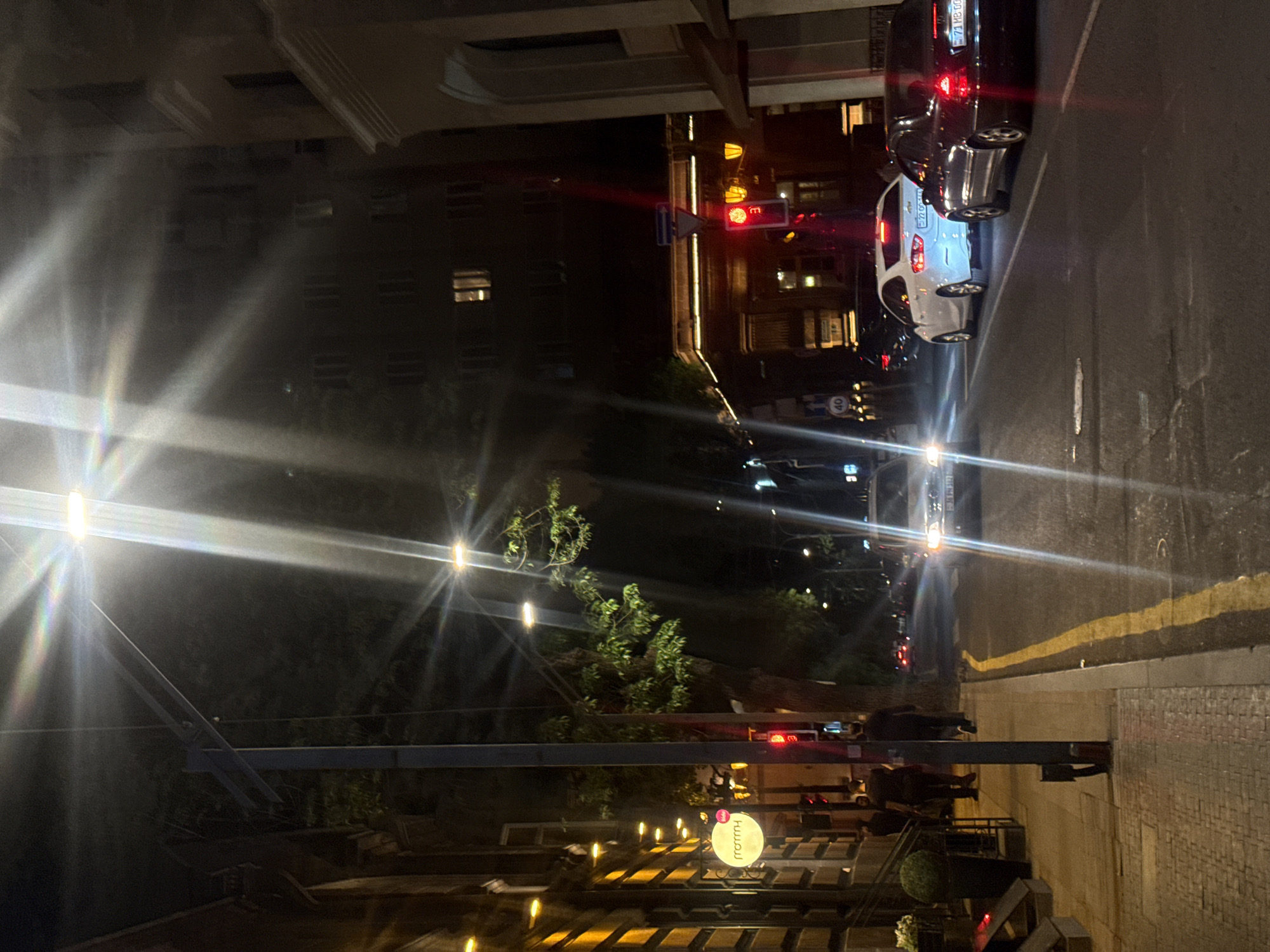 At night in Yerevan, Armenia, cars are parked along a street next to a sidewalk lined with a patio dining area. A restaurant with lit tables, and people are seated. The sky is dark, and streetlights illuminate the scene. A building is visible behind the patio. Traffic lights are visible on the far side of the street.