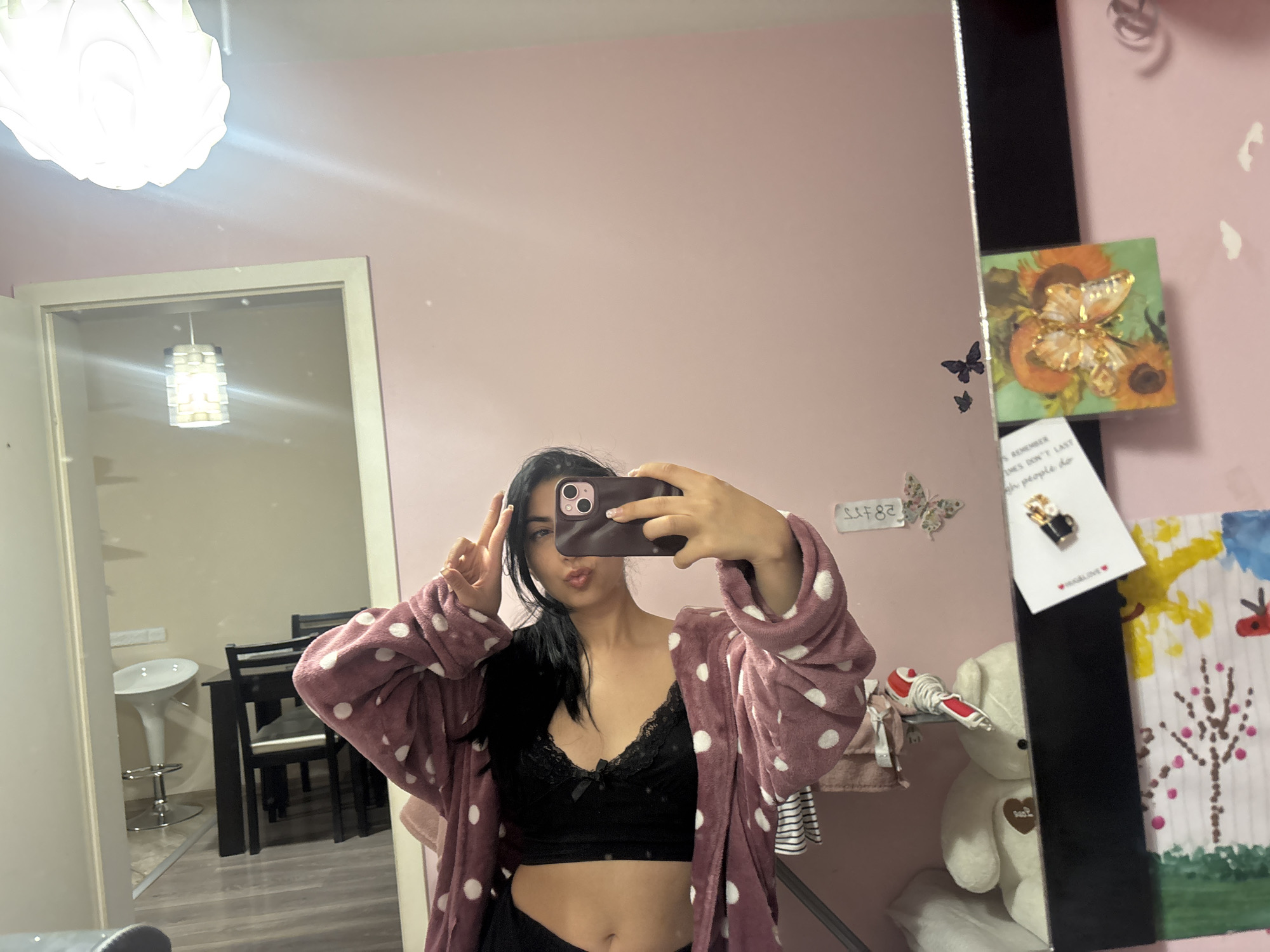 A person stands in front of a mirror, taking a selfie. The individual has dark hair and is wearing a black lacy top and a mauve polka-dot robe. They are holding a phone in one hand and making a peace sign with the other. The background shows a pink wall with decorative elements such as a butterfly sticker, a picture of sunflowers, and a child's drawing. A shelf with a teddy bear and other items is also visible. The reflection in the mirror reveals a dining area with a table, chairs, and a breakfast bar, suggesting the scene is likely inside an apartment or home. There is no specific information to determine the exact location within Plovdiv, Bulgaria.