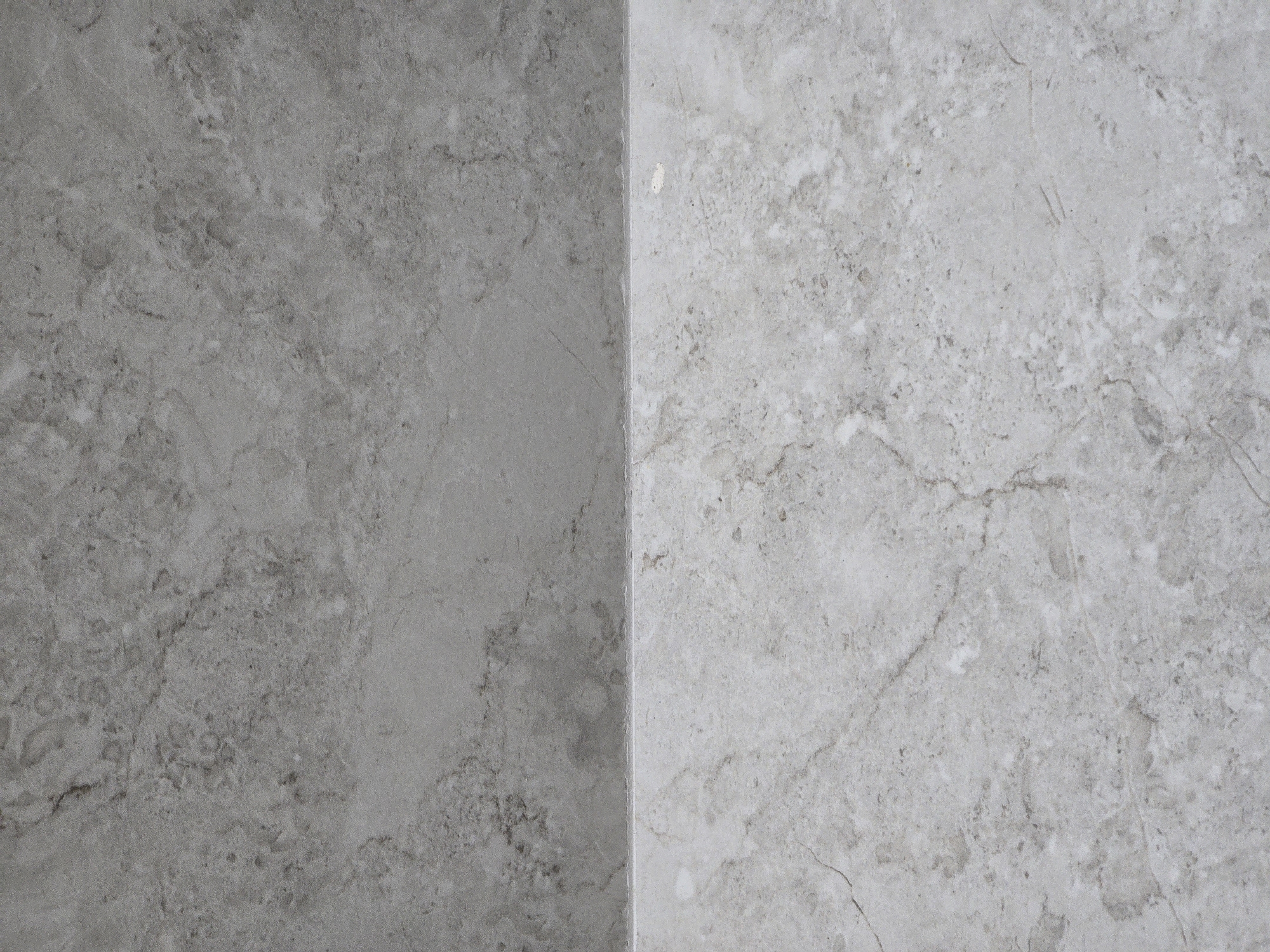 The image shows a close-up view of two tiles, one above the other, separated by a thin horizontal grout line. The upper tile is a darker shade of gray with a marble-like pattern of darker gray veins. The lower tile is a lighter shade of gray, almost white, also with a marble-like pattern of gray veins, some of which appear more pronounced and linear. The surface of both tiles appears to be matte and slightly textured. The lighting is even, highlighting the texture and patterns of the tiles. There are no people, animals, or any other objects visible in the image. The image is a detail shot of building materials. The location of Sütəmurdov, Azerbaijan is irrelevant to the visual content of this image.
