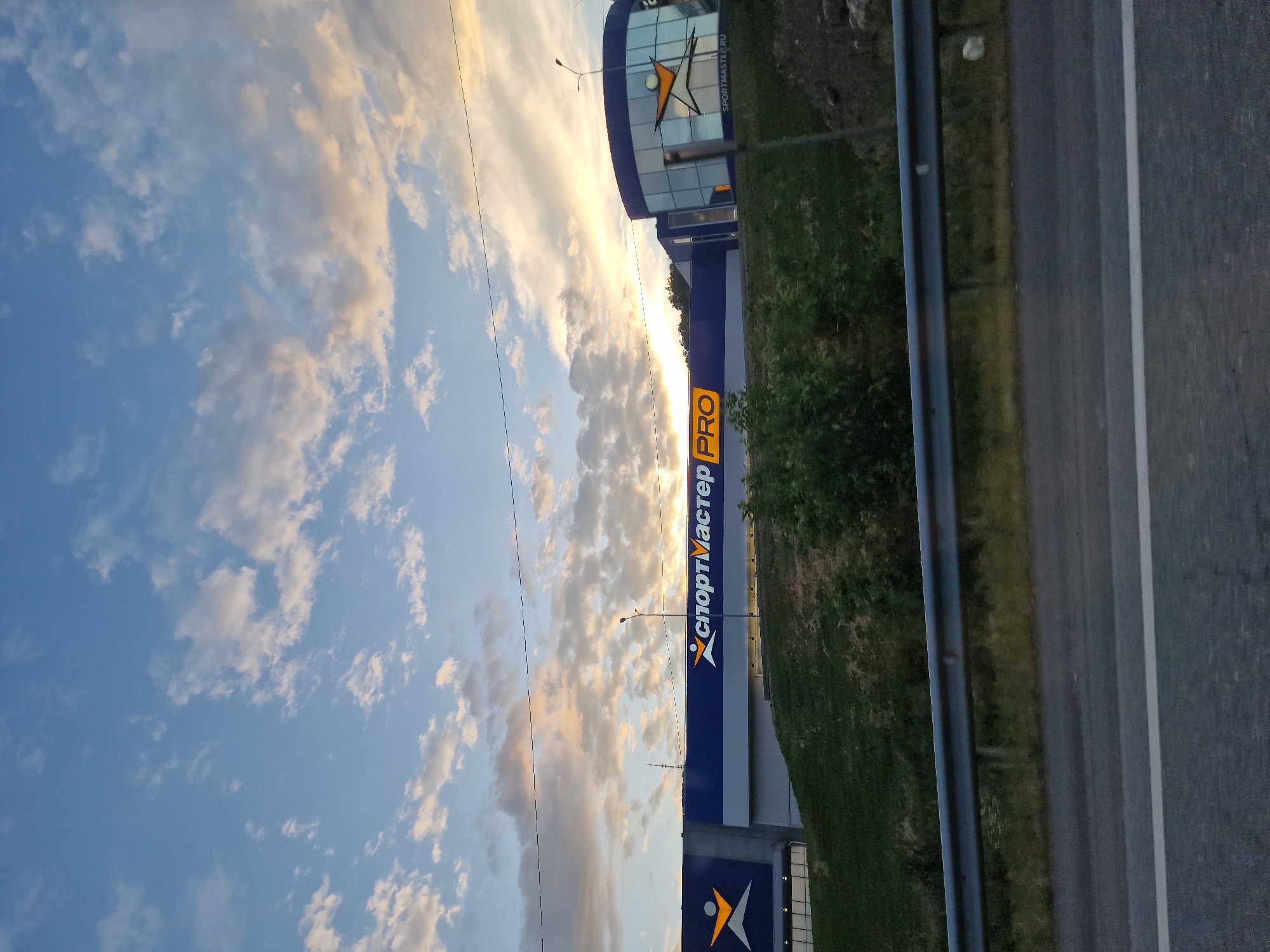 The image captures a wide shot of a Sportmaster PRO store in Goryachevodsky, Russia, under a cloudy sky with hints of sunset colors. The building features a prominent blue facade with large white lettering spelling out "СПОРТМАСТЕР PRO". The store's logo, a stylized orange figure with outstretched arms, is visible on the left side of the building. To the right, a section of the building is rounded with glass windows, also displaying the Sportmaster logo and the website "SPORTMASTER.RU". The store is situated on a grassy incline, with a metal guardrail and a paved road in the foreground. A street lamp is visible on the right side of the building. The overall impression is that of a commercial establishment on the outskirts of a town or city, seen from a road.