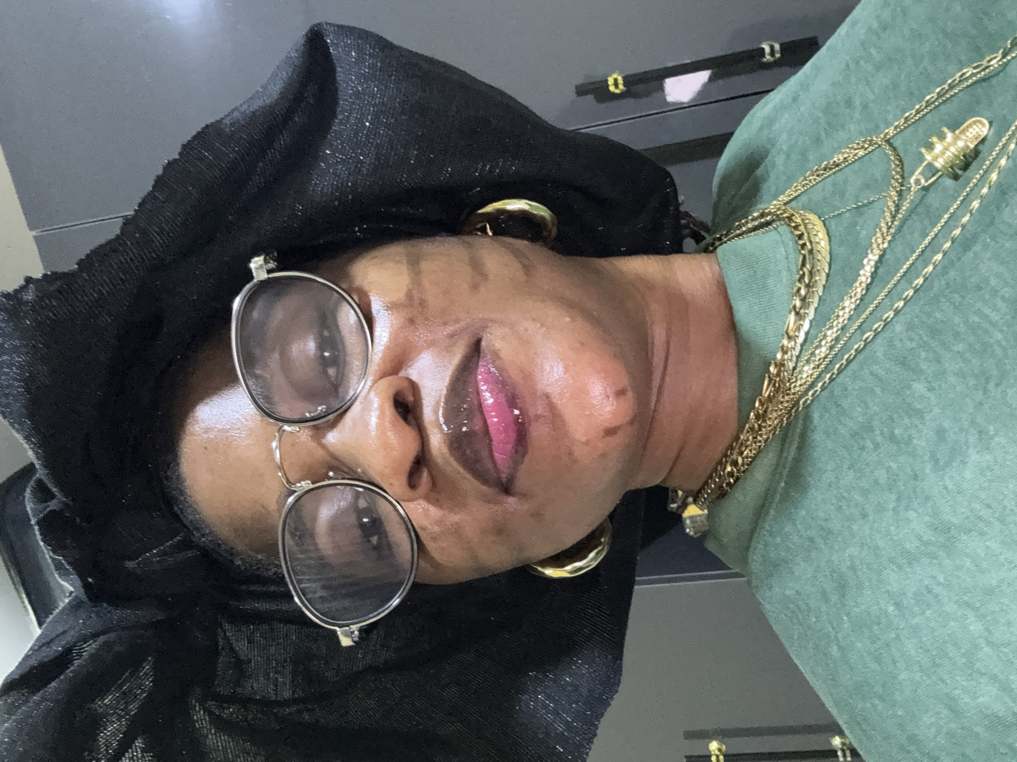 In Ajara, Nigeria, a woman is shown in a close-up, upper-body photograph. She is wearing a black headwrap and round, silver-rimmed glasses. Her lips are glossy and a vibrant pink. She has on a green sweatshirt and is adorned with multiple gold necklaces, including a thick chain and thinner layers with a pendant shaped like a capsule. Large gold hoop earrings are visible on each ear. The background appears to be an indoor setting with grey cabinets and doors.