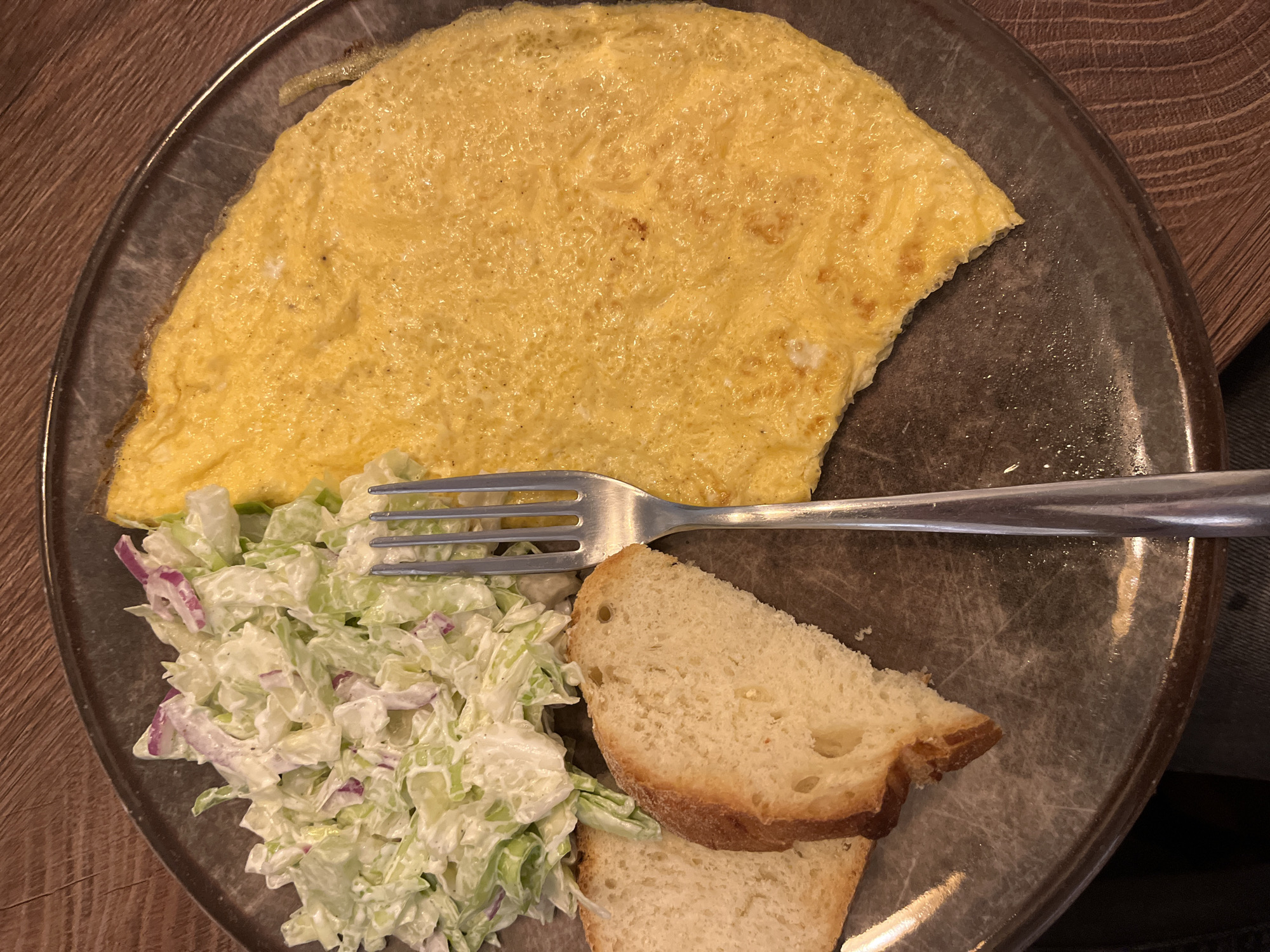 A high-angle shot depicts a plate with an omelet, coleslaw, and two slices of bread. A fork is placed across the plate, overlapping the coleslaw and bread. The scene appears to be indoors, possibly in Budva, Montenegro, though the exact location cannot be determined from the image alone. No people are visible. The focus is on the food items and their arrangement on the plate. The environment is suggested by the wooden table surface visible beneath the plate.