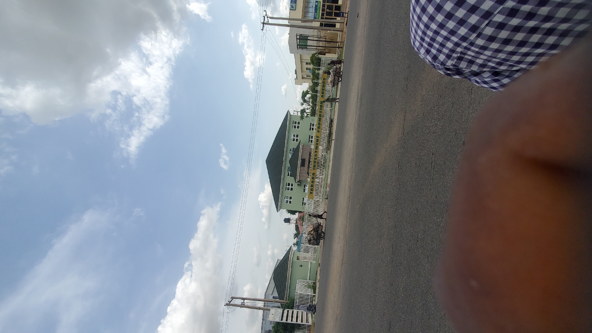 The image captures a street scene in Jalingo, Nigeria. In the foreground, a portion of a person's shoulder and arm is visible, wearing a purple and white checkered shirt. The middle ground features a paved road and a sidewalk or verge lined with a white fence. Beyond the fence, several buildings are visible. A prominent light green building with a dark green roof and numerous windows is situated centrally. To the left of this building is another smaller light green building with a similar green roof. To the right, there is another light-colored building with windows. A person is seen pushing a cart laden with goods on the street. Several other individuals are also present, walking or standing near the buildings. The sky above is partly cloudy with a mix of white and gray clouds against a light blue background. The overall impression is that of a commercial or residential area in a city.