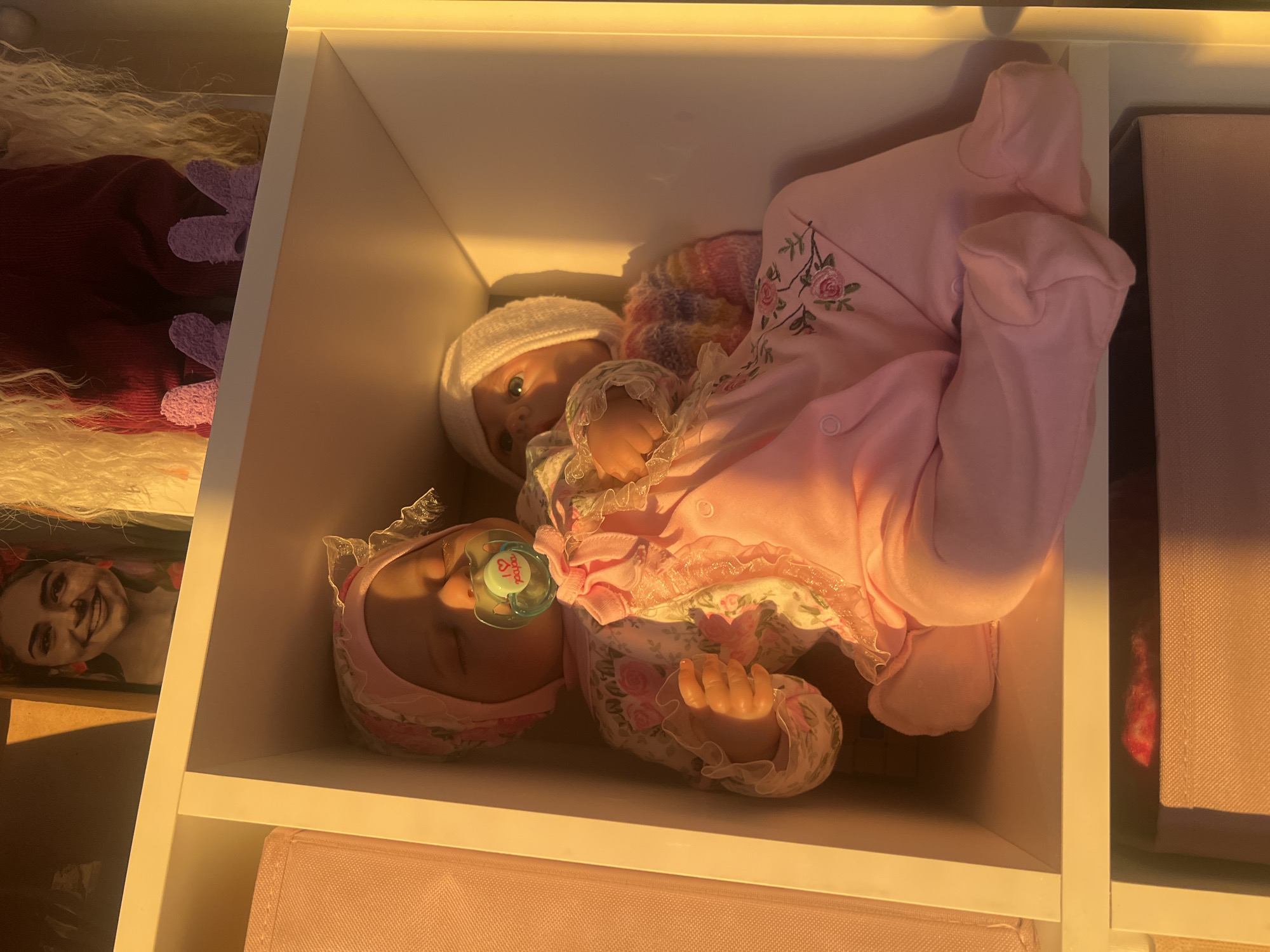 The image depicts two baby dolls inside a white shelving unit. One doll wears a pink floral bonnet with a pacifier in its mouth, while the other doll wears a white cap and a multicolored sweater under a pink floral onesie. The dolls are positioned side-by-side within the shelf. The surrounding environment includes additional items in the shelving unit, such as a box and a photo. There is no information present in the image to suggest this is specifically located in Votkinsk, Russia.