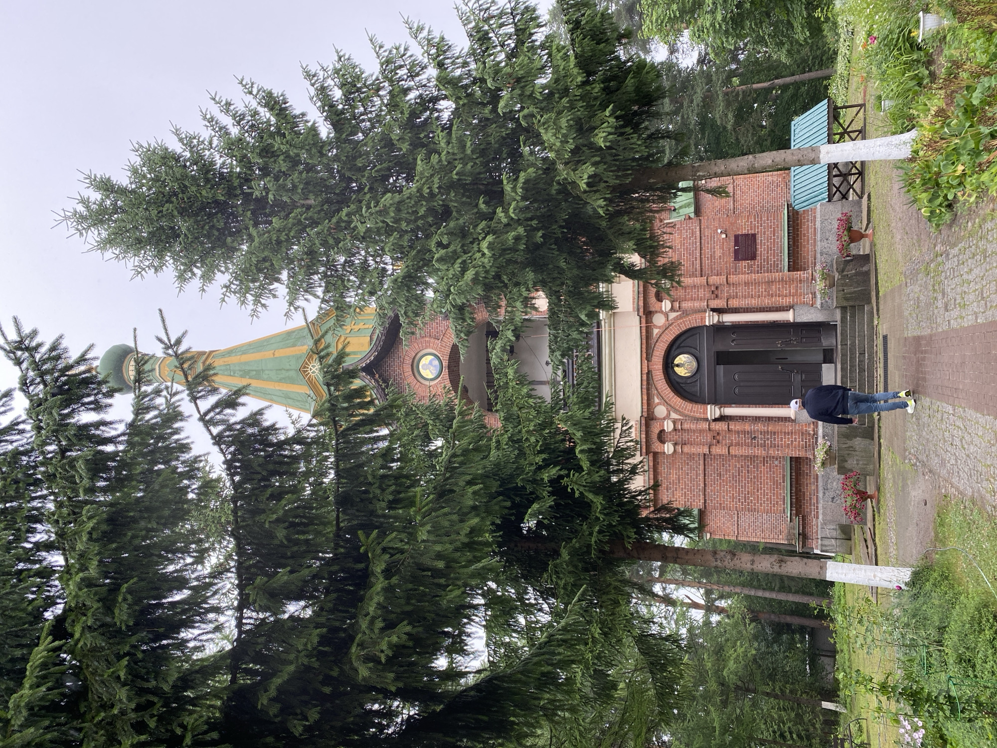 A person walks toward the entrance of the Valaam Monastery Church, also known as the Church of the Resurrection of Christ, located in Priozersk, Russia. The church is constructed of red brick and features a green-domed spire with yellow trim. Two large pine trees frame the front of the building, obscuring some of the facade. The ground in front of the church is paved with cobblestones and brick. A man in a dark jacket and jeans, wearing a white baseball cap, walks away from the viewer towards the church entrance. The sky is overcast. The church appears to be situated in a wooded area.