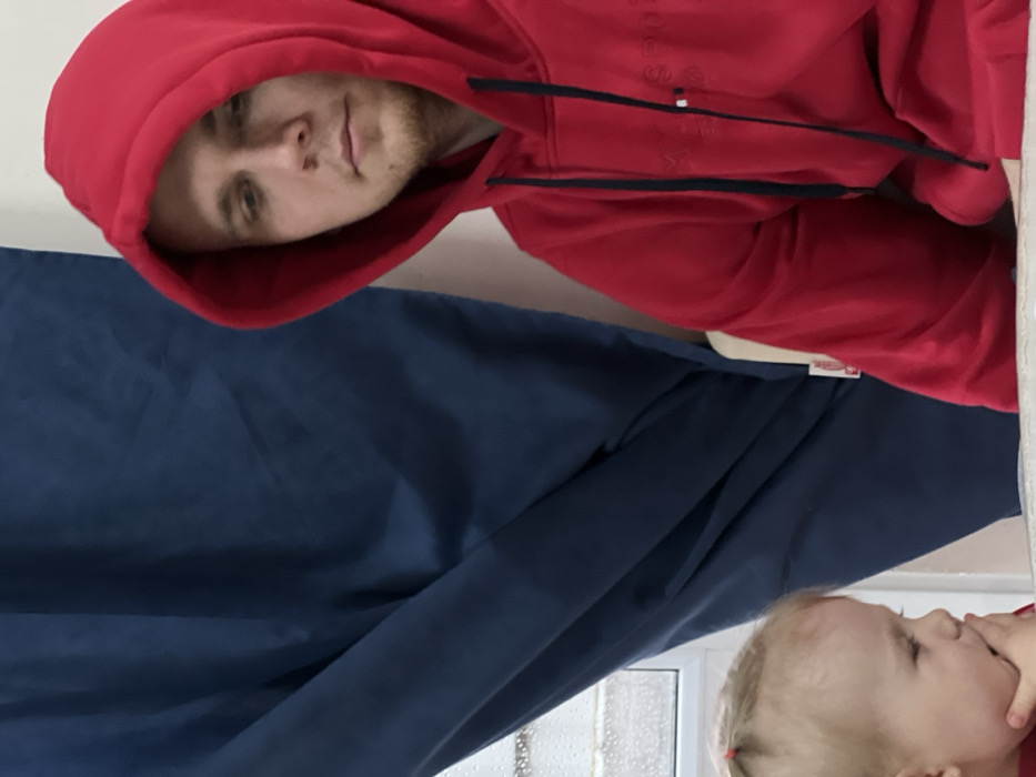An indoor scene depicts two individuals. On the right, an adult male with light-colored hair and facial stubble wears a red hooded sweatshirt with the hood covering his head; he is seated, looking downwards and to the left. In the lower-left corner, a young child with blonde hair tied with two small red clips is partially visible, wearing red clothing and holding her right hand near her mouth; her head is tilted down. A light-colored surface separates the two figures. The background features dark blue curtains partially obscuring a window, through which an exterior view blurred by precipitation on the glass is visible. The wall behind the man is white. The setting is located in the city of Leninakan, Russia.