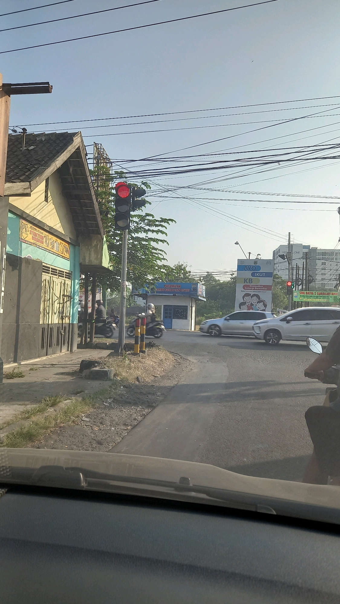 The image captures a street scene in Ngadirejo, Indonesia, focusing on a traffic intersection under a clear, bright sky. A traffic light, illuminated with a red signal, is prominently positioned on the right side of the frame. Several vehicles are present, including two motorcycles with riders on the left side of the road, one silver sedan in the middle, and a silver SUV to its right, all seemingly stopped or moving slowly due to the red light. A portion of a building with a yellow facade and a tiled roof is visible on the far left. Further down the road, a small kiosk with blue and white signage is situated. Overhead, a complex network of electrical wires crisscrosses the sky. The foreground shows the interior of a vehicle, with a dashboard and windshield wiper visible. The overall impression is a typical urban street view in Indonesia, with traffic, infrastructure, and daily life unfolding at an intersection.