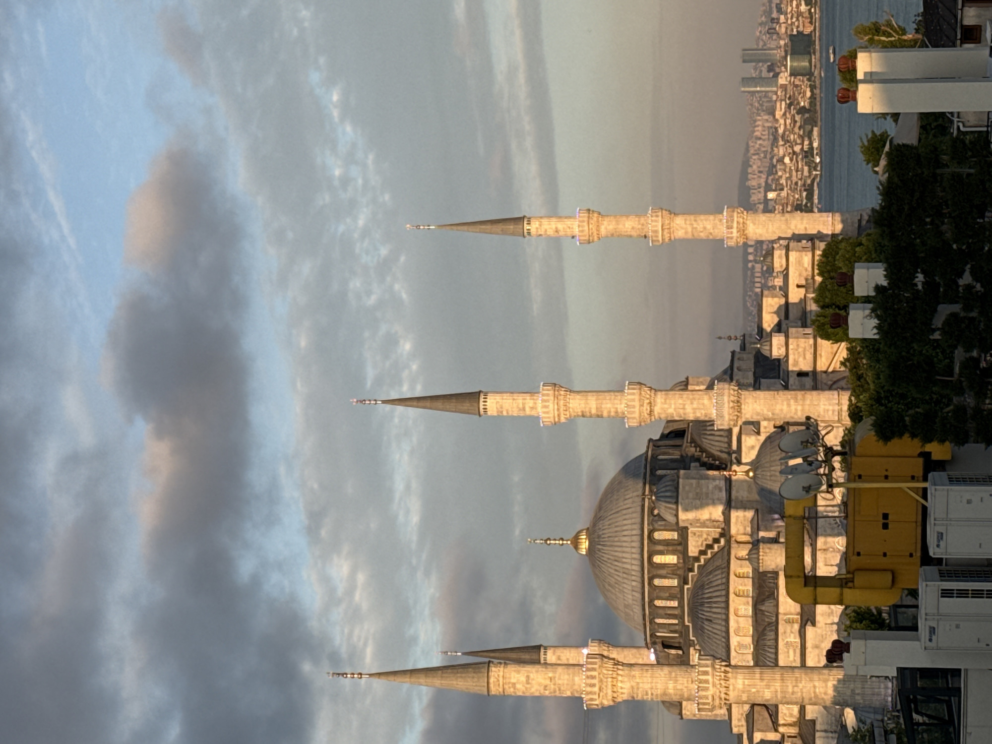 The image depicts the Hagia Sophia in Istanbul, Turkey, at what appears to be sunrise or sunset, given the warm, golden light illuminating the facade and minarets. The grand structure, characterized by its massive dome and multiple slender minarets, dominates the central part of the frame. The architectural details of the mosque, including arched windows and intricate stonework, are visible. In the foreground, various rooftop elements are present, including what appears to be an industrial generator, air conditioning units, and white chimneys with red crowns. Beyond the mosque, a body of water, likely the Bosphorus, stretches across the mid-ground, with the cityscape of Istanbul visible on the opposite shore. The skyline features a mix of older buildings and modern skyscrapers, including twin towers. The sky above is partly cloudy, with diffused sunlight creating a soft, atmospheric effect. No people are visible in the image.
