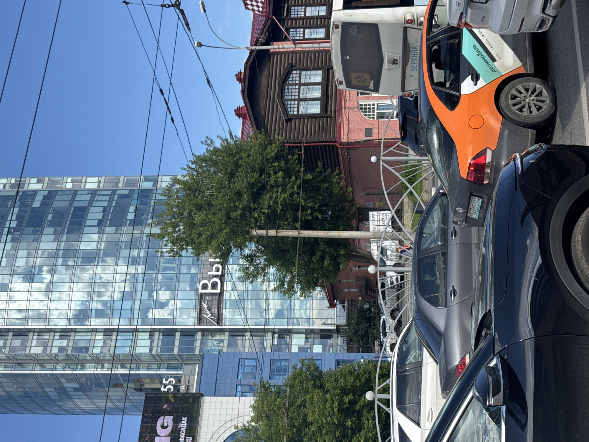 The image depicts a street scene in Yekaterinburg, Russia, under a clear blue sky, suggesting it's daytime and likely fair weather. The perspective is from a street level looking upwards and diagonally, with a significant portion of the frame filled with vehicles and buildings.
In the foreground, several cars are parked or driving. A dark-colored car is prominent on the right, with its side and rear visible. Next to it is a silver-grey sedan, and further back, an orange and dark grey compact car with a distinctive graphic design on its side. Behind these, parts of a white bus or delivery vehicle are visible.
The background features a mix of architectural styles. On the left, a modern glass-fronted skyscraper reflects the sky and clouds, with a white sign on its facade showing what appears to be Cyrillic text, including "Bb". Below this, another sign displays "55". To the right of the skyscraper is a traditional wooden building with dark-trimmed windows and a red roof, contrasting sharply with the modern structure. Adjacent to this is a brick building. Trees with lush green foliage are interspersed between the buildings, indicating a potentially urban setting with green spaces.
Overhead, numerous power lines crisscross the sky, a common feature in many urban environments. A metal decorative fence or railing is visible in the middle ground, separating the street from what might be a sidewalk or park area. The overall impression is a bustling urban environment with a mix of old and new architecture, and daily life continuing with cars and pedestrian activity (though few people are clearly visible). The presence of Cyrillic text provides a strong indicator of the Russian location.