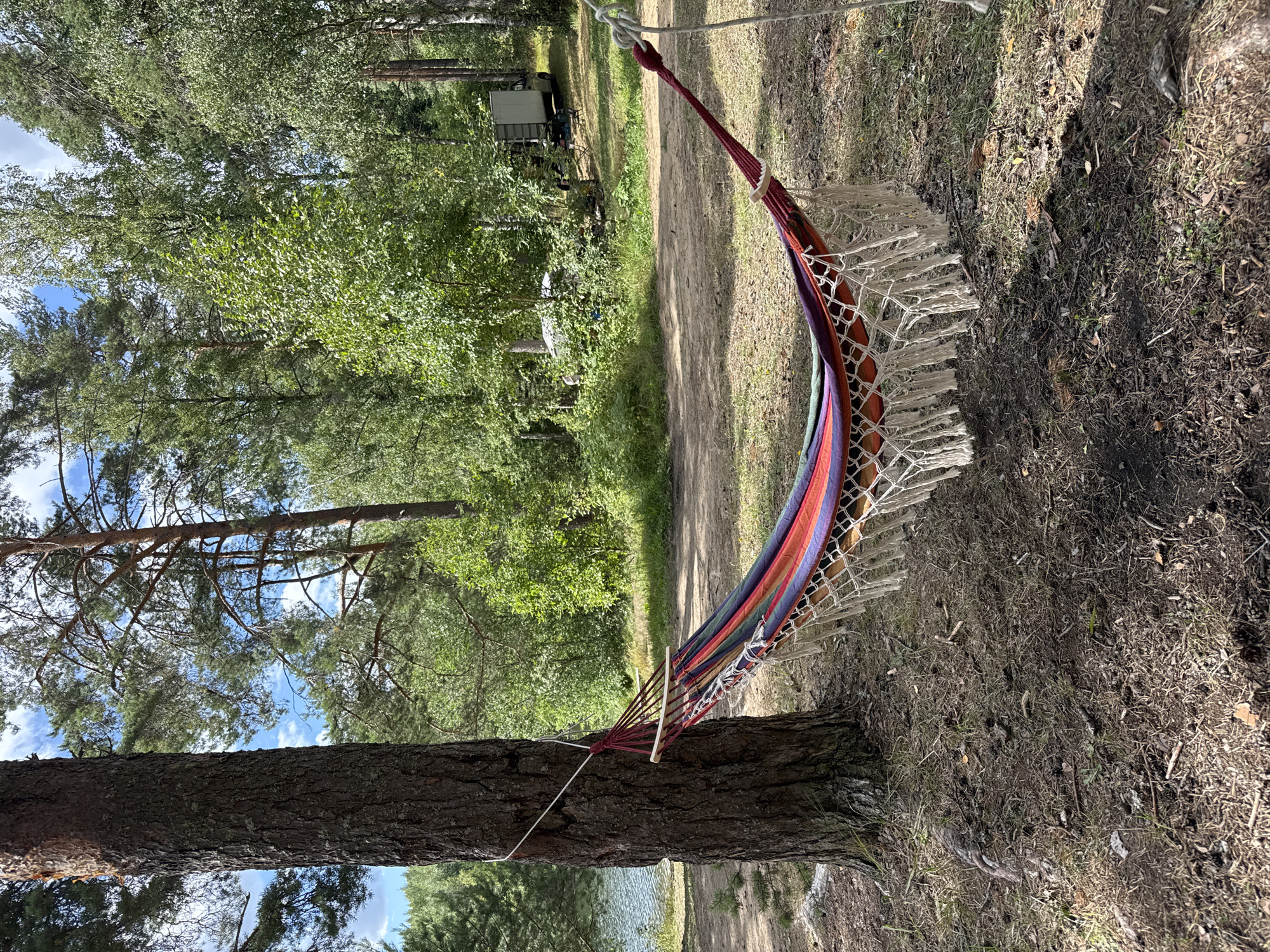 A striped hammock is suspended between a large pine tree and another unseen anchor point, with the scene set in a wooded area near a body of water, possibly Lake Ladoga, in Lumivaara, Russia. The hammock, featuring vibrant red, blue, green, and orange stripes, is adorned with white macrame fringe. The ground beneath is covered in pine needles, dry leaves, and scattered pinecones, indicating a natural, possibly remote, location. In the background, dense green foliage of various trees dominates the landscape, with a glimpse of a lake to the left. Further in the distance, a small white caravan or camper is visible, suggesting a recreational or camping setting. The sky above is partly cloudy, with patches of blue sky peeking through the tree canopy. No people are visible in the image, and the overall impression is one of peaceful solitude.