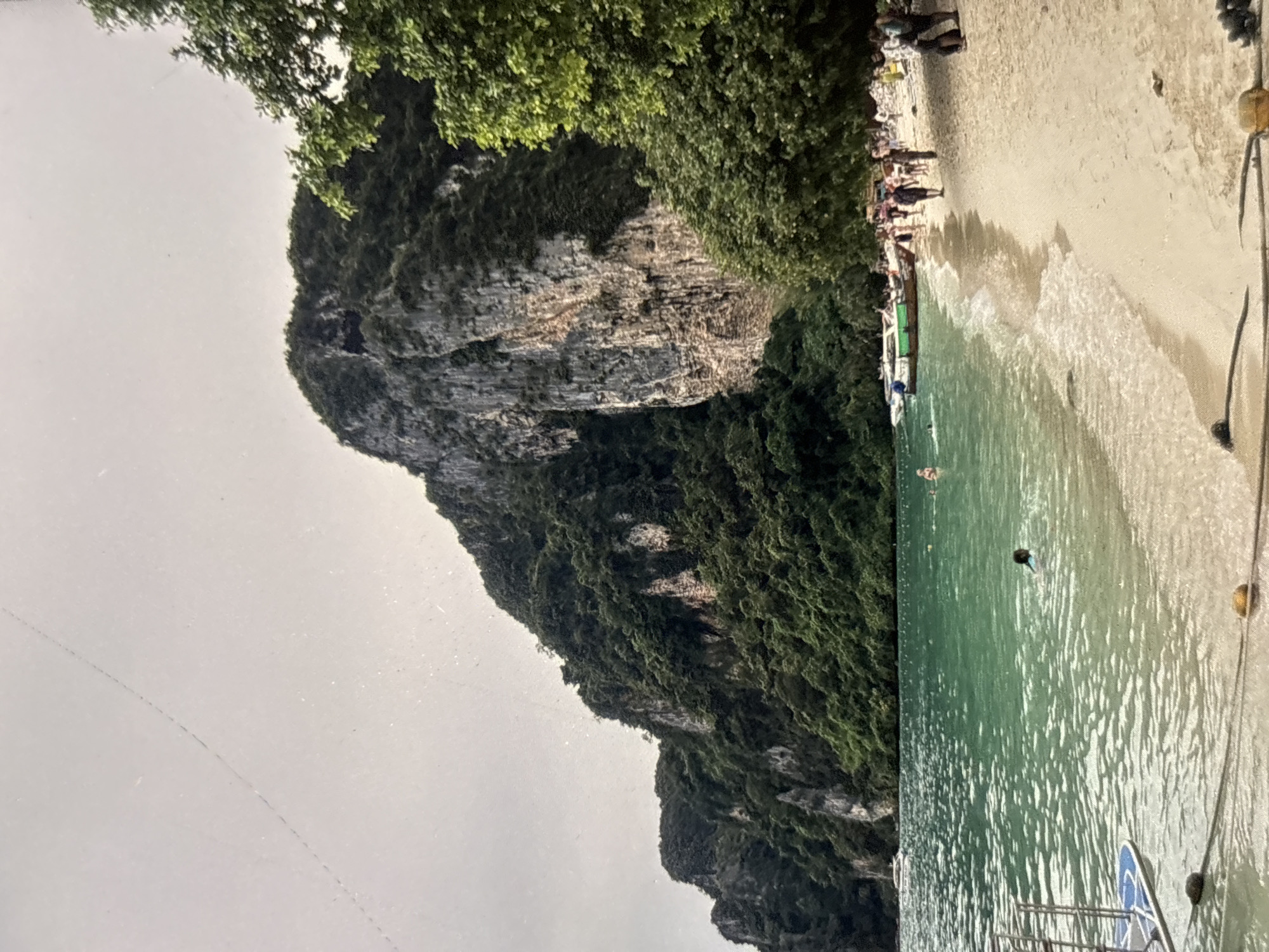 This image depicts a tropical beach scene in Chalong, Thailand, with a towering, lushly vegetated limestone cliff dominating the background. The turquoise water is clear, revealing a sandy bottom near the shore where a person is swimming. Further out, several boats, including a longtail boat, are moored in the calm sea. A sandy beach lines the water's edge, with a group of people walking and standing on it. Trees with green foliage partially obscure the view of the cliff from the right side of the frame. The sky is overcast, creating a diffused light.