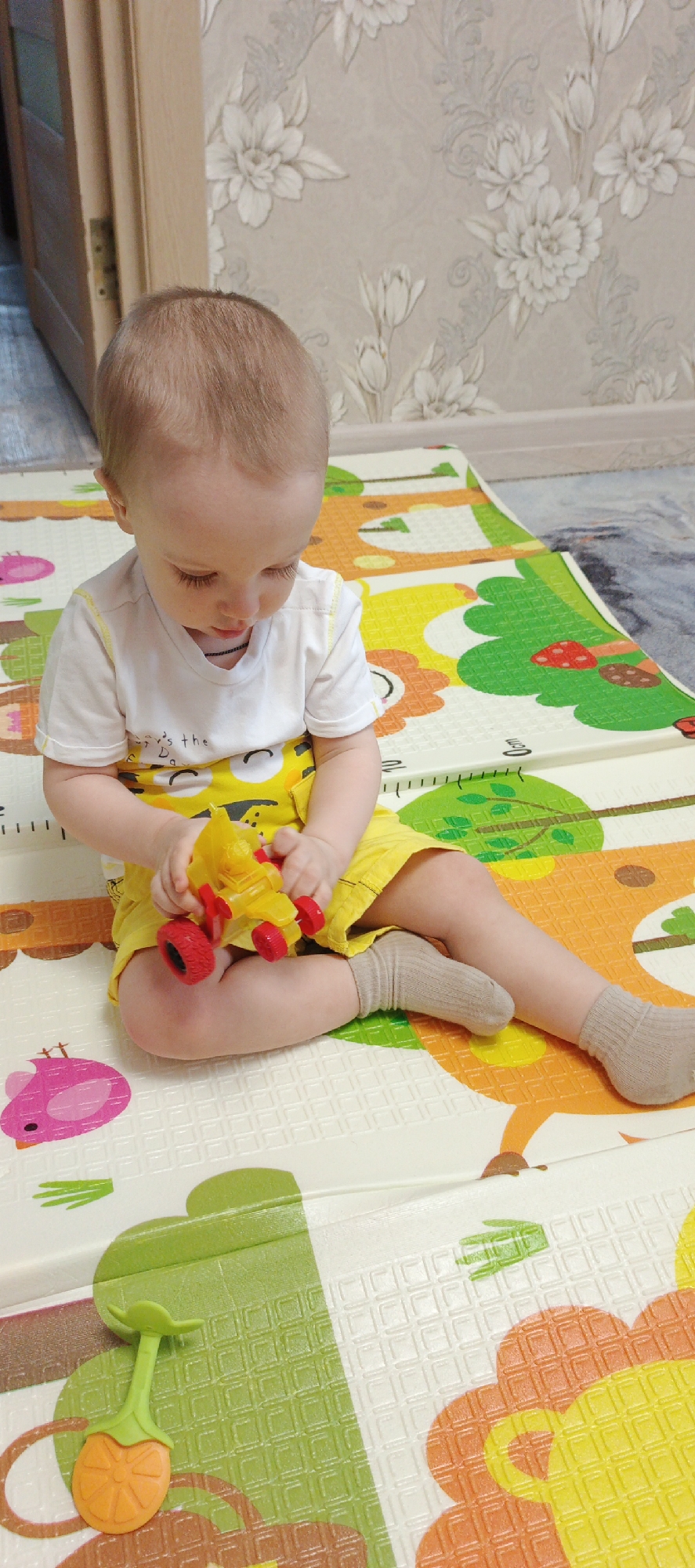 A child is sitting on a colorful play mat with a yellow toy car in their hands. The child is wearing a white t-shirt with a yellow graphic and yellow shorts. They are sitting cross-legged, looking down at the car. The play mat has cartoon animals, trees, and other designs on it. The background includes a floral patterned wall and a partially visible door. The location is Chaikovsky, Russia.