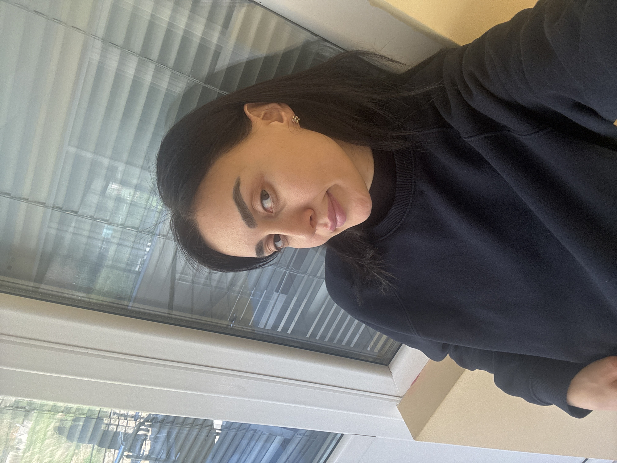 A woman with dark hair and a dark sweatshirt is sitting indoors. She is near a window with blinds. Outside the window, trees and buildings are visible. The location cannot be determined as Tbilisi, Georgia from visual information alone.