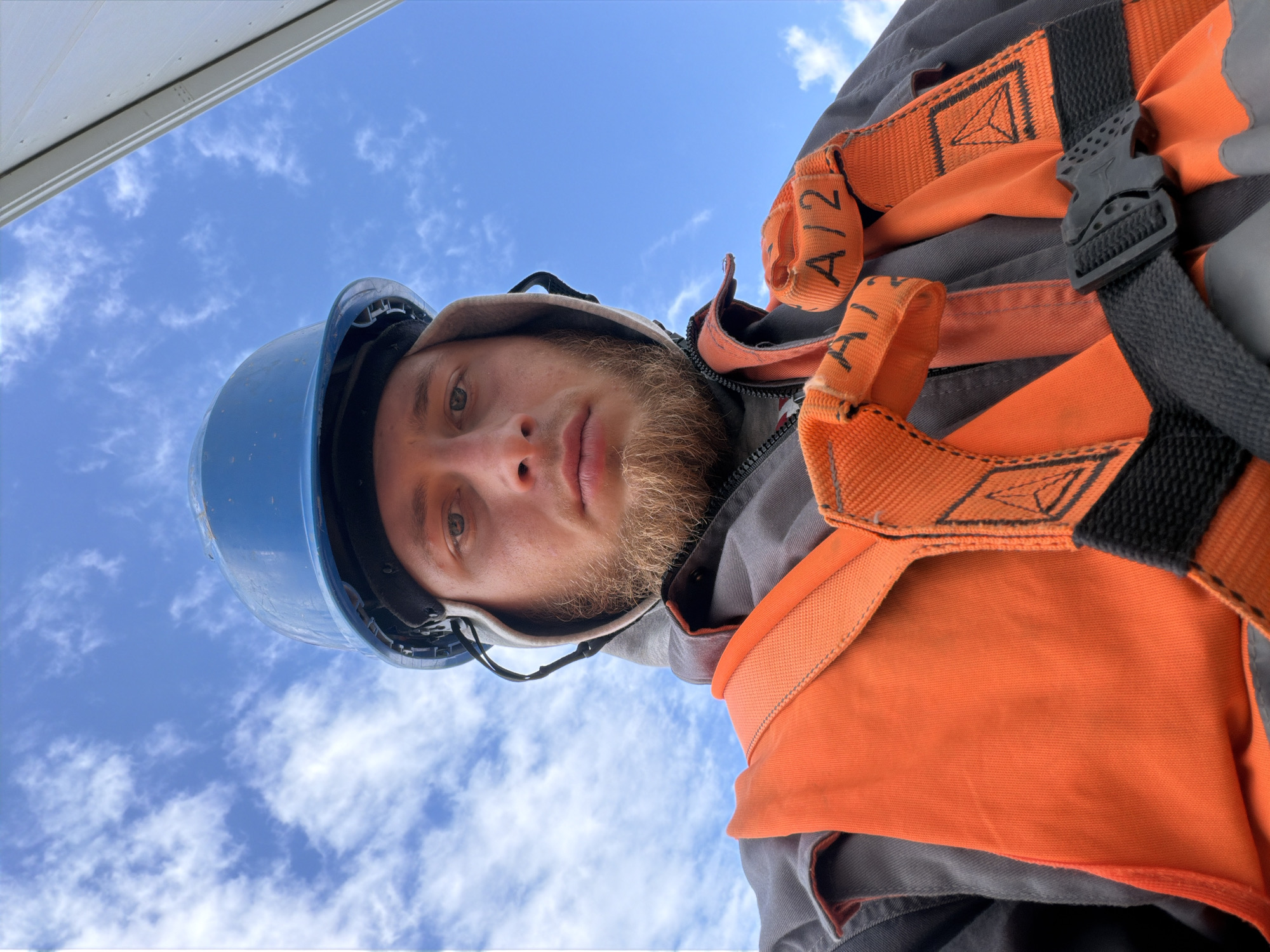 In the city of Stróża, Poland, a close-up, low-angle, selfie-style photograph captures a young Caucasian male against a bright blue sky with scattered white clouds. The man is wearing a blue hard hat and a grey hooded jacket, over which is a bright orange safety vest and harness. The harness straps are black and orange, with some lettering visible on the orange straps, appearing to be "A12". He has a light brown beard and is looking directly at the camera with a neutral expression. The background is predominantly the sky, with a sliver of a white structure visible in the upper right corner.