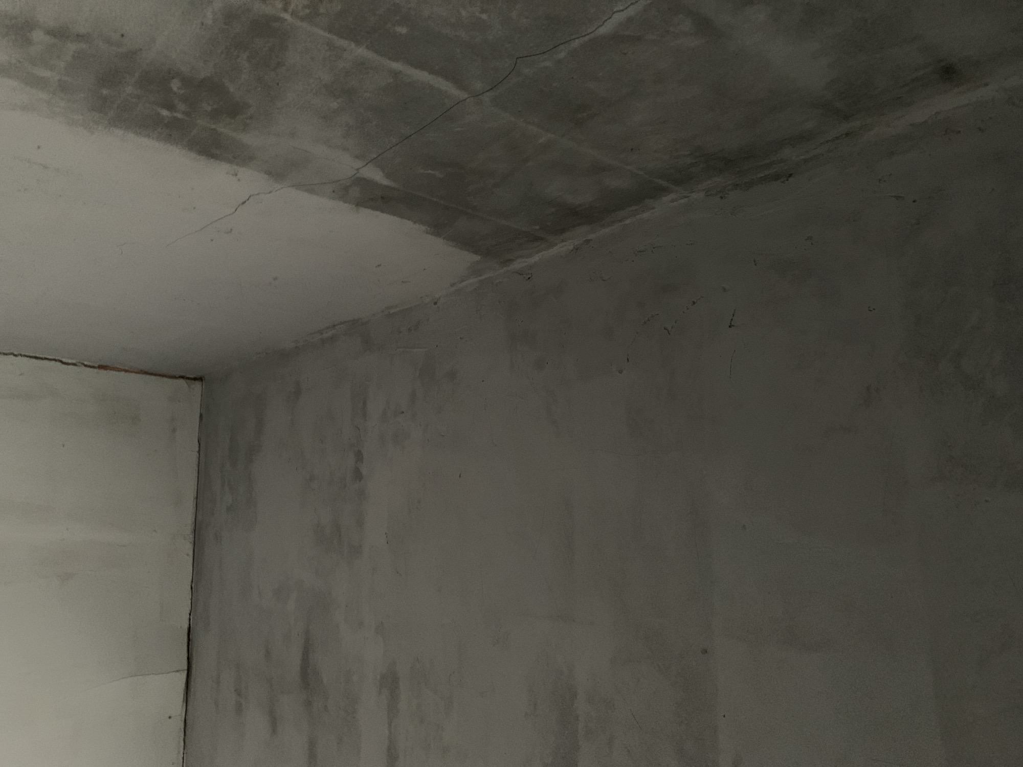 The image shows an interior corner of an empty room with unfinished concrete walls and floor. The walls appear to be made of cinder blocks with some plastering. There are visible cracks in the plaster and evidence of water stains or discoloration on the wall. The floor is also concrete and appears to be roughly finished. The lighting is dim, casting long shadows. There are no people or objects in the room. This scene is not located in the city of Utama, Indonesia.