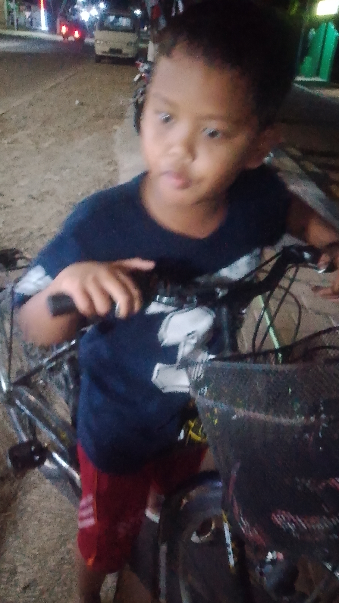 A young boy with dark hair and brown eyes is standing next to a bicycle on a street at night. He is wearing a dark blue t-shirt and red shorts. The bicycle has a basket on the front. In the background, there are streetlights and vehicles, including a white van. The ground appears to be paved with dirt or gravel. The image was taken in Karanglangu, Indonesia.