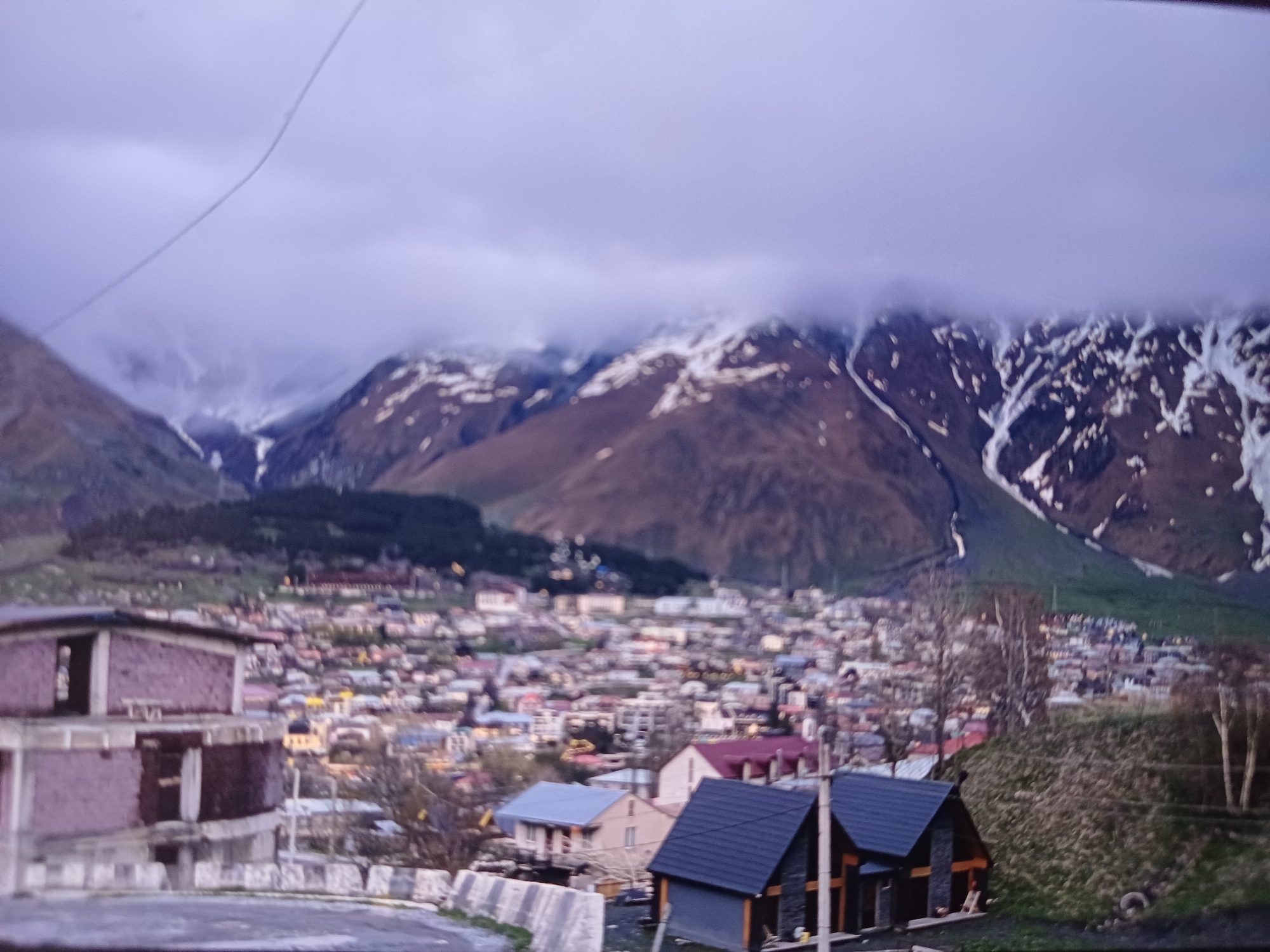 The image shows a high-angle view of the town of Stepantsminda, Georgia, situated in a valley between mountains. In the foreground on the left, there are several modern-style wooden houses with dark roofs. Beyond these houses, the town extends into the distance, characterized by a dense collection of buildings with colorful roofs, some of which appear to be churches or other prominent structures. A river or road runs through the town, with trees and greenery interspersed. To the right, the mountainous landscape dominates the scene, with steep slopes covered in patches of snow and rock. A thick layer of clouds partially obscures the upper portions of the mountains, creating a dramatic atmosphere. The overall impression is one of a remote, mountainous settlement during a transitional season, likely late autumn or early spring, given the mix of bare trees and snow.