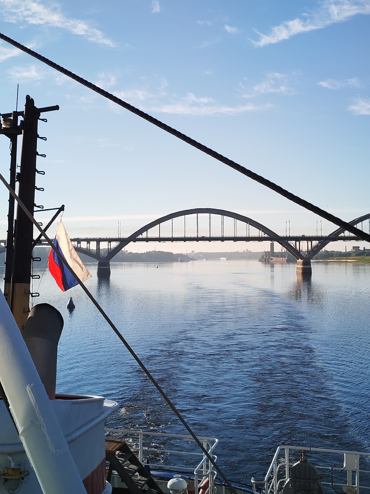 The image captures a view from a boat on a wide river, likely under a clear sky with scattered clouds. A large arched bridge spans the river in the distance. On the boat in the foreground, a Russian flag is prominently displayed, attached to a mast. The wake of the boat is visible on the water behind it, indicating movement. The water appears calm and reflects the sky and the bridge. The overall scene suggests a peaceful journey on the river.