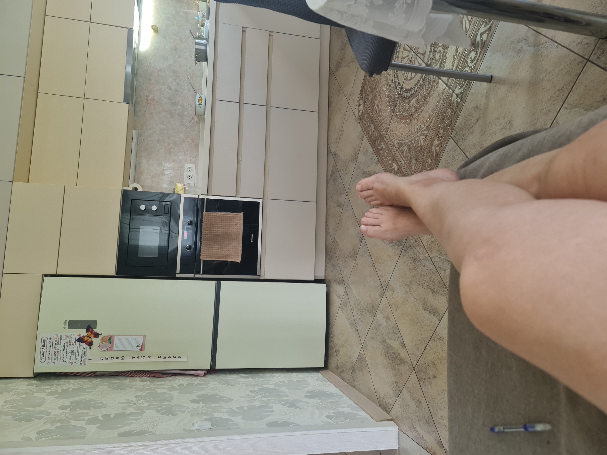 The image depicts a first-person perspective of a person resting in what appears to be a kitchen area. The floor is tiled with large, light brown square tiles arranged diagonally. The person's bare feet and legs are visible in the foreground, crossed at the ankles, resting on a grey upholstered surface. To the left, a portion of a wall with a light-colored patterned wallpaper is visible. Adjacent to the wallpaper is a cream-colored refrigerator with a magnetic butterfly and several handwritten notes attached to its door. A microwave is integrated into the cabinetry above a built-in oven. The kitchen cabinetry is a pale beige color. A patterned rug is visible on the floor between the person's feet and a dark grey chair. The overall environment suggests a residential kitchen.