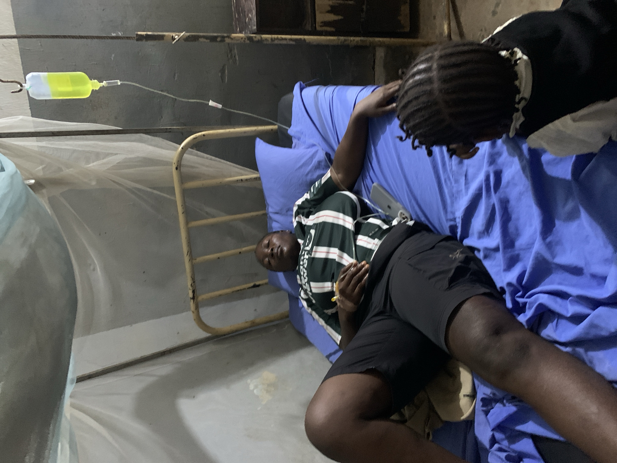 A photograph shows a man lying in a hospital bed under a mosquito net in Akwanga, Nigeria. The man has an intravenous drip connected to his arm. Another person is standing next to the bed, looking at the man. The bed has blue sheets, and the room appears to be sparsely furnished. The image is taken from a high angle, looking down on the scene.