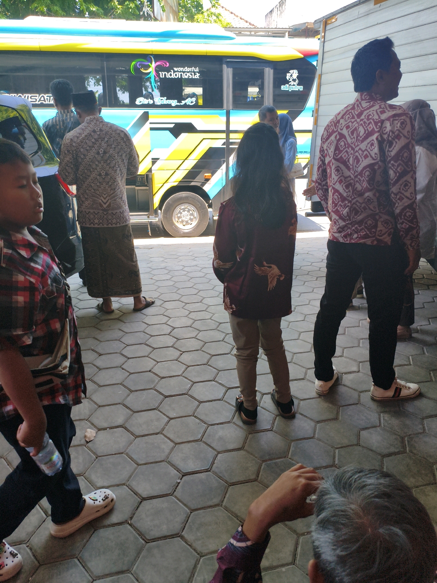 In the city of Kuripan, Indonesia, a group of people are gathered near a brightly colored bus. The bus is yellow and blue with "Wonderful Indonesia" and a stylized Indonesian flag logo visible on its side. Several individuals, including men, women, and children, are standing on a hexagonal tiled pathway in front of the bus. Some of the men are wearing traditional Indonesian attire, such as batik shirts and sarongs. A young boy in a red and black plaid shirt is holding a bottle of water. The scene appears to be taking place outdoors, possibly at a bus station or a tourist stop. The lighting suggests it is daytime, with sunlight casting shadows on the ground.