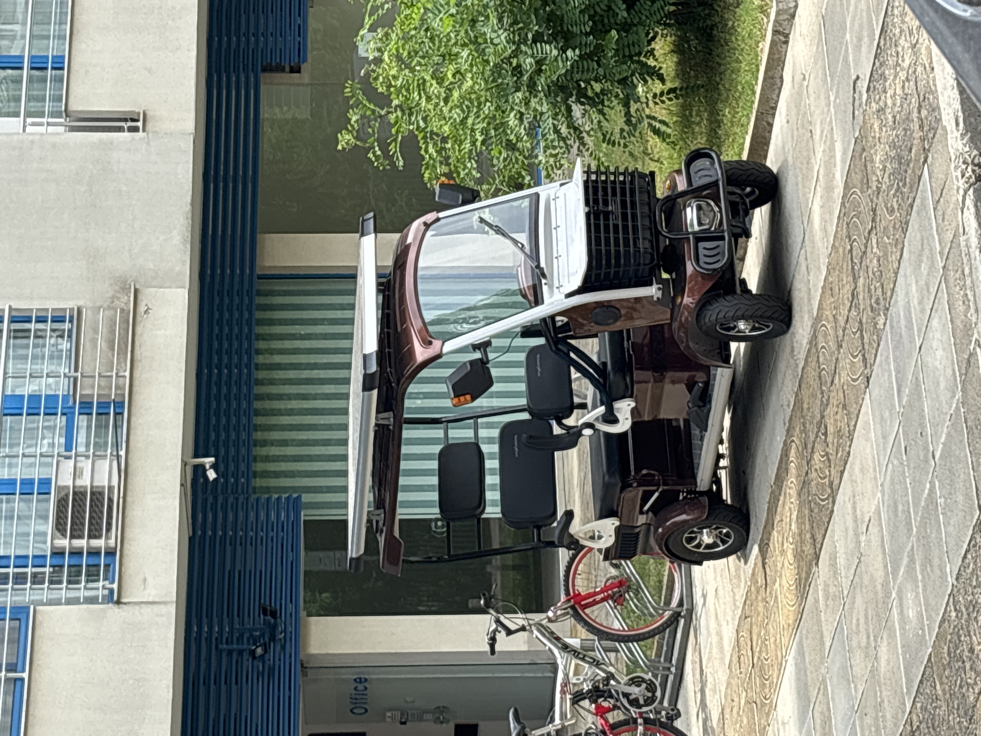 The image shows a brown and white three-wheeled electric scooter parked on a tiled sidewalk in front of a building. The scooter has a canopy roof, a basket on the front, and two seats. To the left of the scooter, there is a bicycle. The building appears to be an office building with a blue striped facade and several windows. There is also a green bush to the right of the scooter. The scene suggests a typical urban environment in Nesebar, Bulgaria.