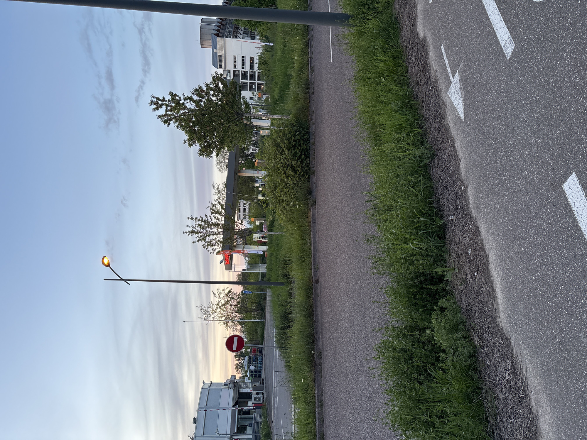 The image depicts a view along a road in Strasbourg, France. The road is marked with white arrows. To the left of the road, there is a sidewalk and a grassy area with bushes and trees. In the background, buildings, a gas station with the "Total" brand visible, and street signs, including a "do not enter" sign, are visible. A street light is on, casting a warm glow. The sky is a mix of blue and pastel colors, suggesting either sunrise or sunset. The buildings appear to be modern. There are no people visible in the image.