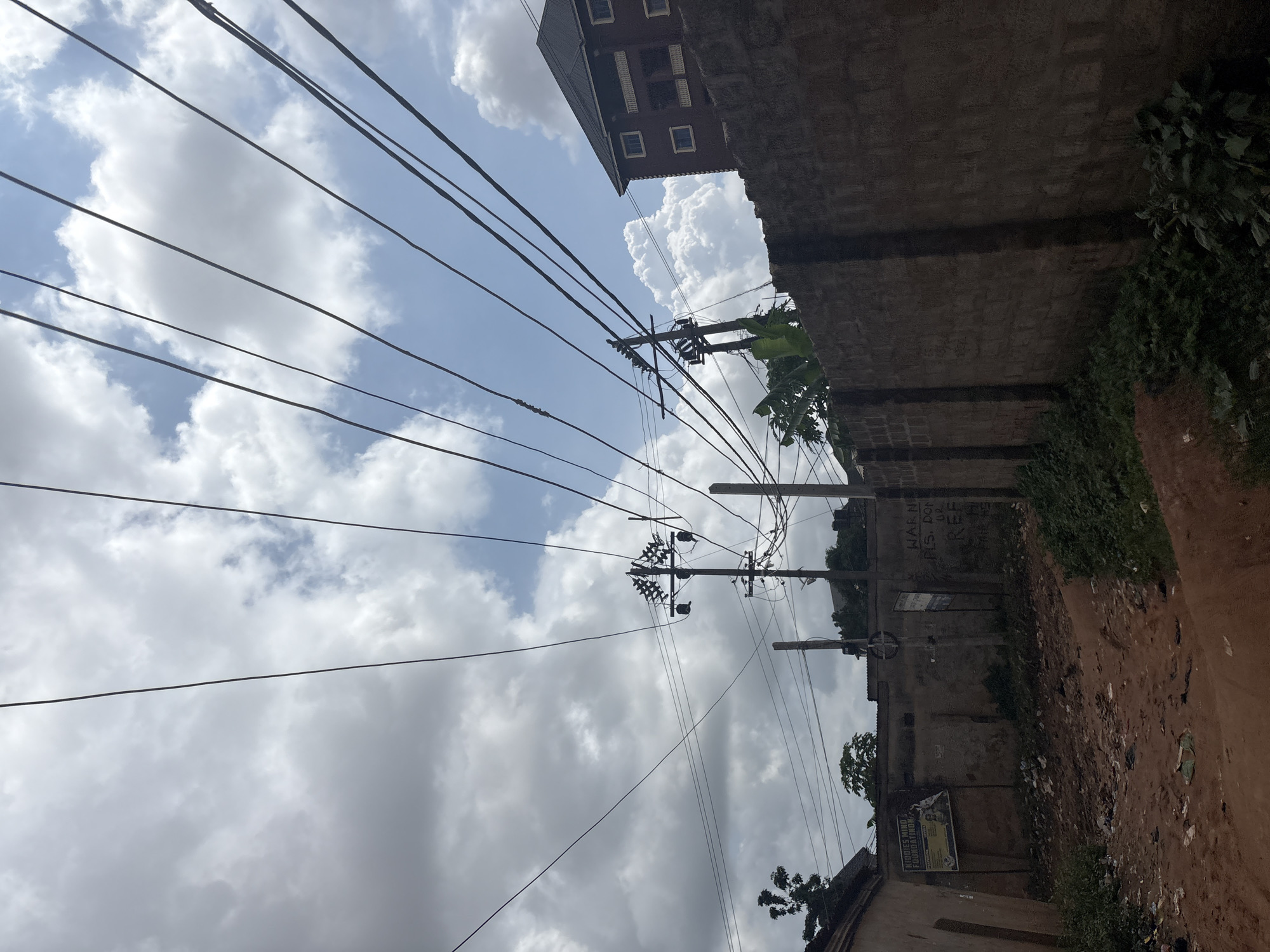 The image depicts an outdoor scene with a focus on a network of overhead power lines against a sky filled with clouds. The perspective is tilted, looking upwards from a street-level view. The power lines stretch diagonally across the frame, supported by utility poles.
To the right of the image, a brick wall runs along the edge of the scene, with some patches of overgrown green vegetation. Above the wall, a building with a dark roof and visible windows can be seen. Further down the wall, some graffiti is present, including the words "WARN" and "FIS. DON'T OR REE". Another poster is affixed to the wall, displaying text that is partially legible, with the words "KNIGHTS MIND FOUNDATION" clearly visible.
The ground visible at the bottom right appears to be a dirt path with scattered debris and some trash. The overall impression is of a somewhat unkempt urban or semi-urban environment. The sky suggests it might be daytime, possibly late morning or afternoon, given the bright lighting and distinct cloud formations. The weather appears to be partly cloudy, with a mix of sunlight breaking through the clouds. There are no people visible in the frame, and the scene itself doesn't indicate any specific ongoing activity or event, presenting more of a static street view. The location cues of Nkpor, Nigeria are noted in the prompt, but not directly inferable from visual elements alone, other than a general sense of a tropical or sub-tropical climate suggested by the vegetation.