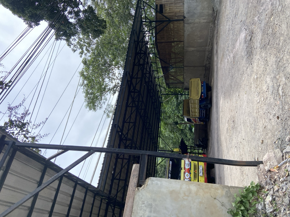 The image shows a dirt lot in Soreang, Indonesia, with a metal roof overhead. The coordinates of the location are -7.042 latitude and 107.498 longitude. The lot is covered with dirt and small stones. A yellow dump truck is parked in the middle of the lot. There is another truck visible in the image, partially obscured. There are a number of electrical wires and cables overhead. The area surrounding the lot is covered in trees, including a bamboo structure, and there is a concrete wall that partially encloses the lot. The image is captured at a slightly oblique angle from the edge of the lot, looking towards the parked dump truck. The overall scene appears to be that of a construction site.