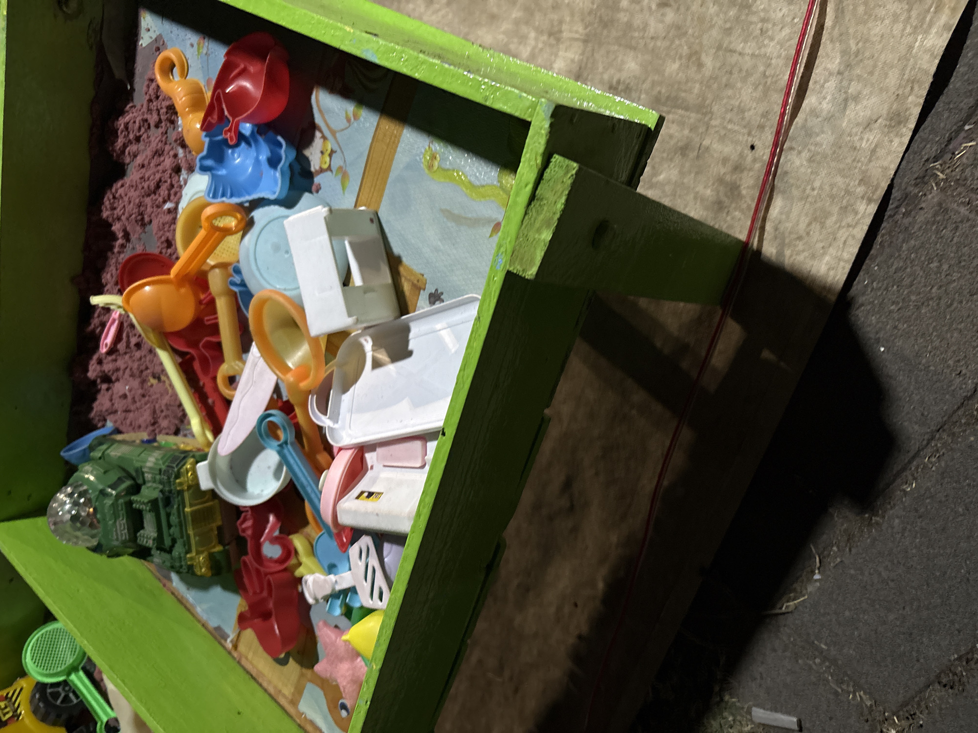 The image shows a green wooden sandbox filled with toys and kinetic sand. Among the toys are a green toy truck with a shiny dome, various colored plastic shovels and molds, a yellow cone, and pink and white plastic containers. The sandbox is situated outdoors on a paved surface, likely a yard or patio. The overall scene is consistent with a children's play area. The location is Palimanan, Indonesia, and the time of day appears to be evening or night, given the lighting and shadows.