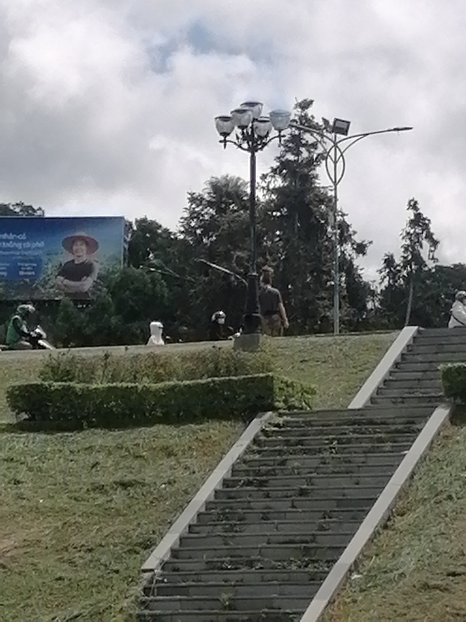 The image is a daytime shot of a city park in Da Lat, Vietnam. A set of concrete stairs ascends a grassy hill, with a trimmed hedge running parallel to the steps. Debris is scattered on the stairs. On the hill's crest, several figures are present. One person stands near a black lamppost. Another individual sits on the stairs. Further to the left, other people are situated near a billboard featuring a person wearing a traditional Vietnamese hat. The sky is overcast with scattered clouds.