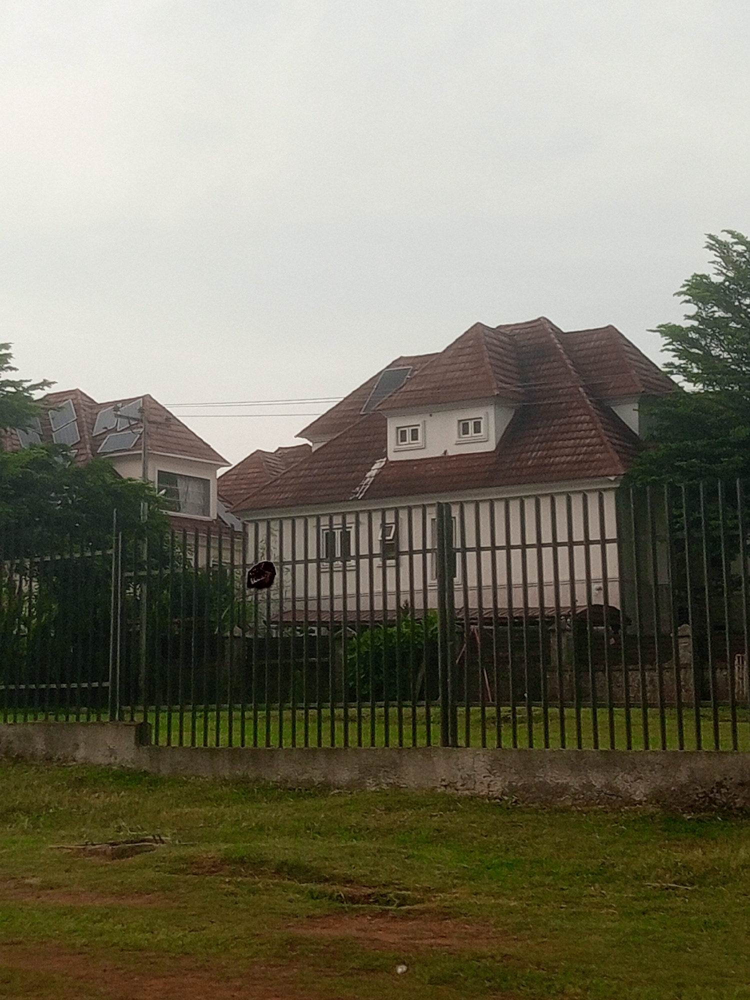 The image shows two large houses with red tiled roofs, one partially obscured by trees on the left and the other more prominently displayed on the right. Both houses have multiple dormer windows. The house on the left has several solar panels installed on its roof. A tall, dark metal fence with vertical bars runs across the foreground, separating the viewer from the houses and their grounds. Behind the fence, there is lush green vegetation. The foreground consists of a patch of dry grass and dirt. The sky is overcast and a pale grey. The scene appears to be located in Gwarinpa, Nigeria, based on the provided context. There are no visible people in the image.